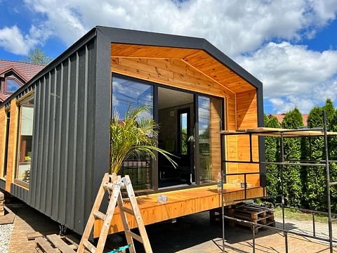 Nowoczesny, mobilny dom z czarną elewacją i drewnianym wykończeniem, częściowo na paletach, z palmą w doniczce przy wejściu i rozstawioną drabiną, na tle błękitnego nieba i zieleni.