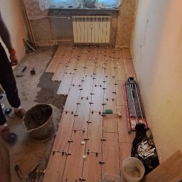 Filip Niećko - Układanie podłogi z płytek imitujących drewno w pokoju z oknem, widoczne krzyżyki dystansowe, wiadro z zaprawą, narzędzia glazurnicze oraz częściowo ułożone płytki.