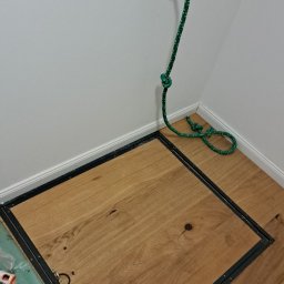 Filip Niećko - Zamknięty właz podłogowy z drewnianą klapą obramowaną czarną metalową ramą, w rogu pomieszczenia, z zieloną liną przymocowaną do ściany.