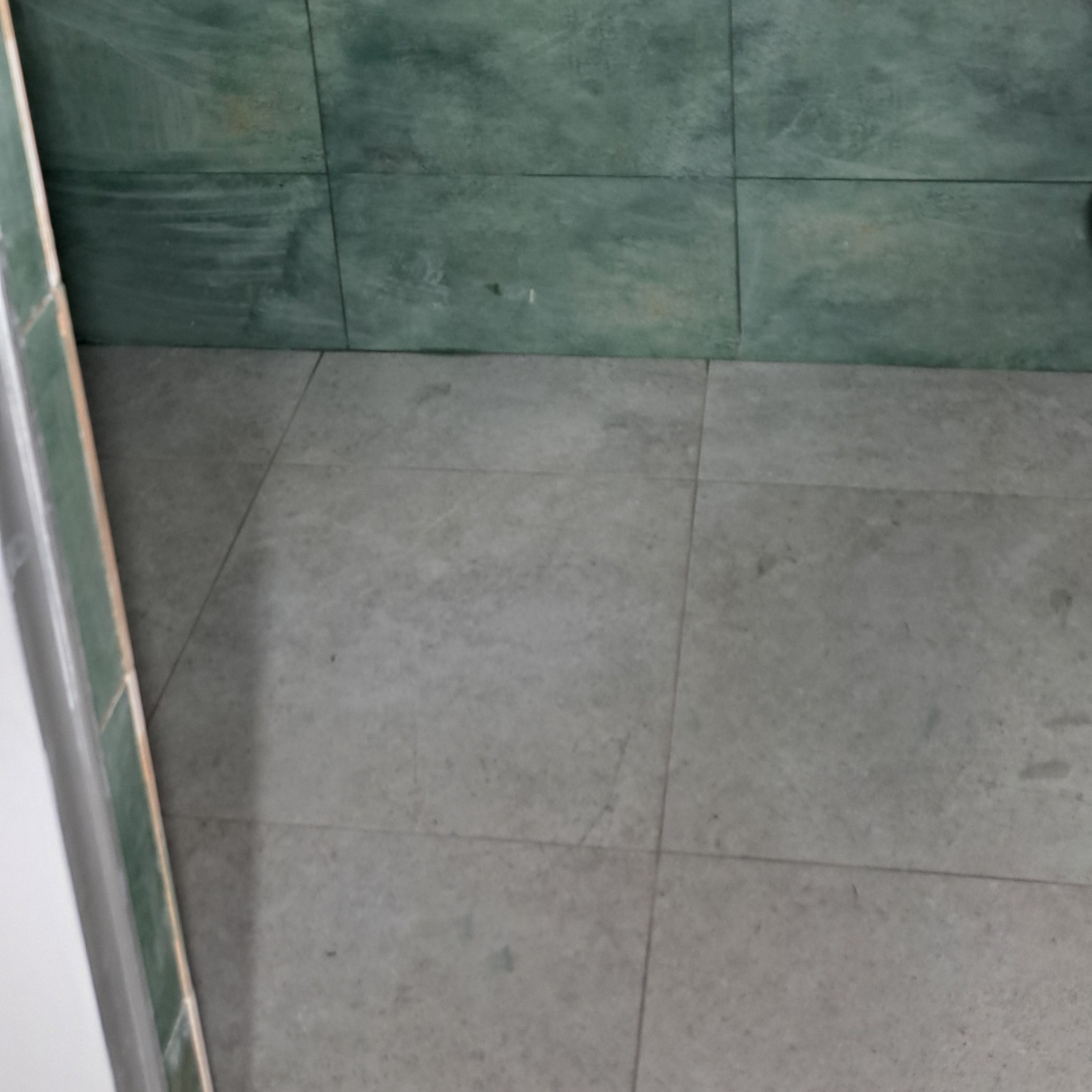 Szare płytki podłogowe w łazience z zieloną glazurą na ścianach, widoczne fugi i fragment białej ramy drzwiowej.