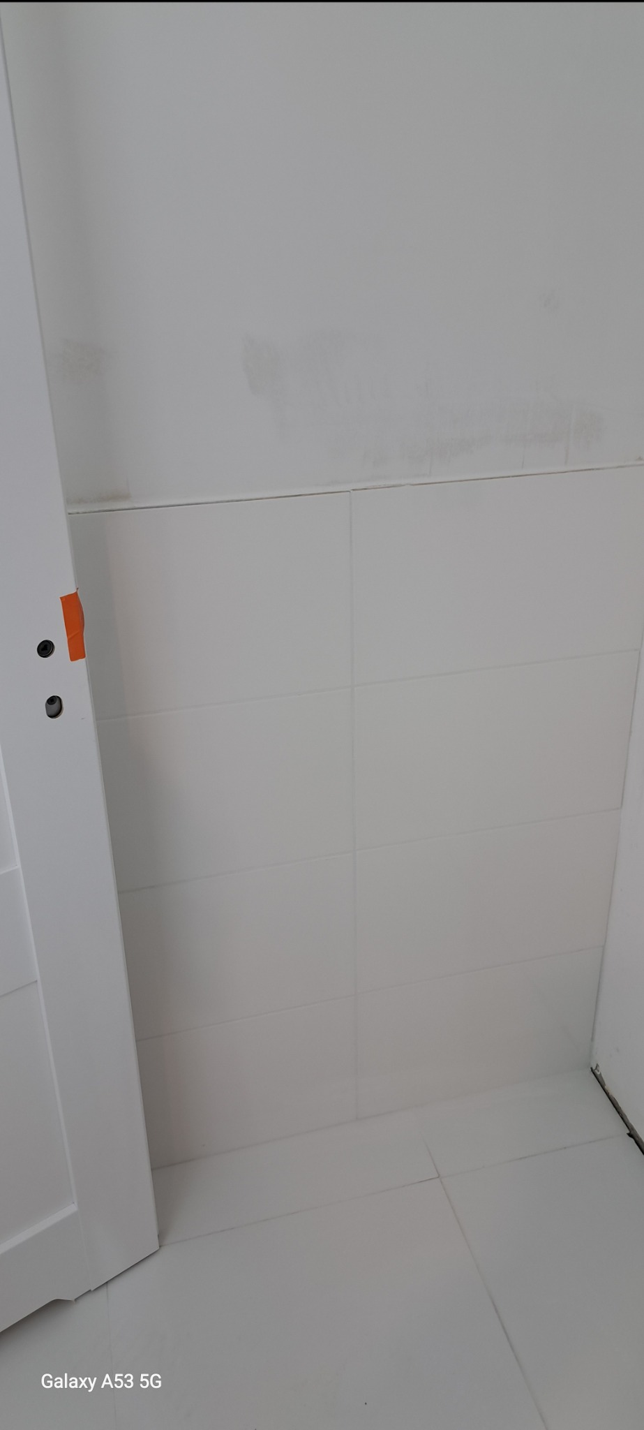 Biała, częściowo otwarta ościeżnica drzwiowa z pomarańczową taśmą zabezpieczającą, na tle białych, prostokątnych płytek ściennych i podłogowych w łazience, widoczne ślady po remoncie na suficie.