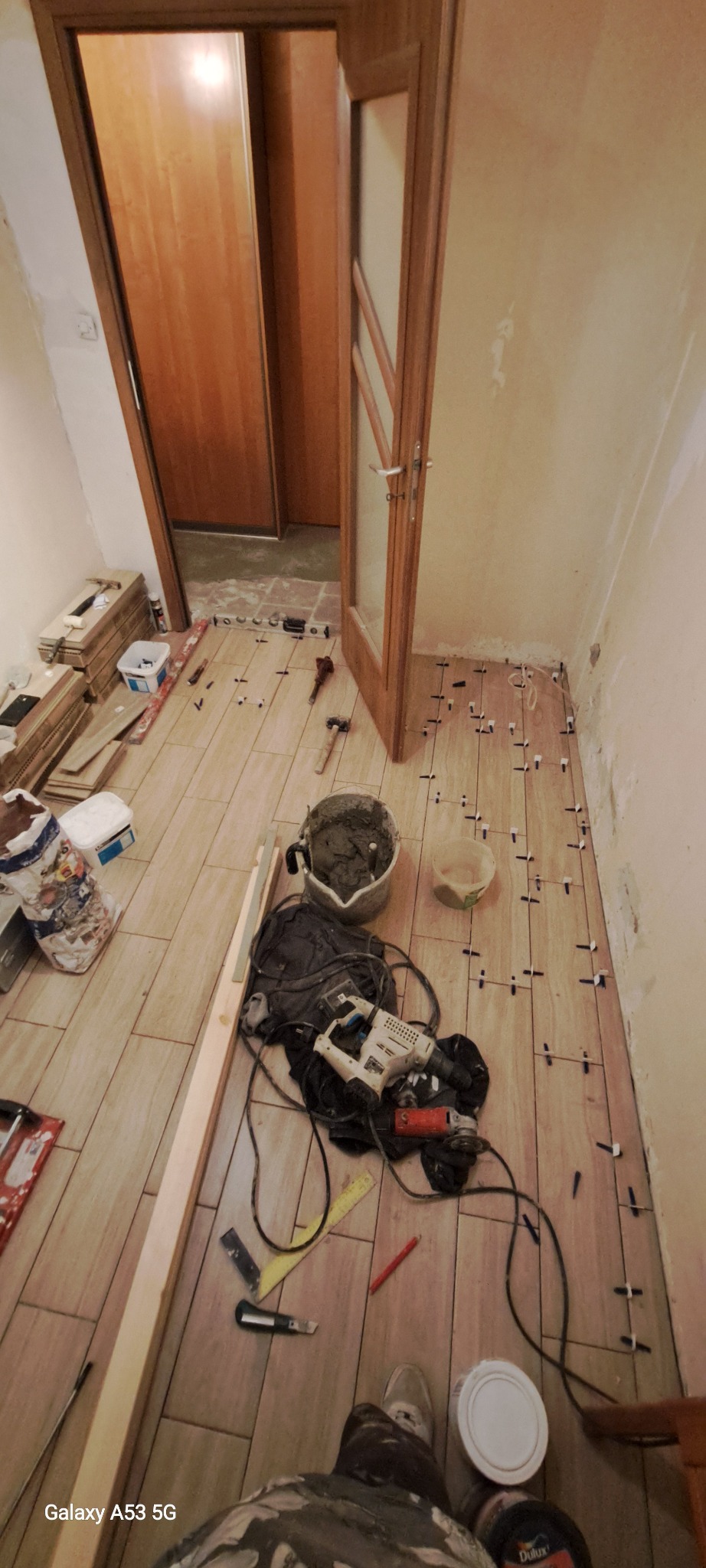 Układanie podłogi z płytek imitujących drewno w korytarzu, widoczne narzędzia: poziomica, szpachelka, wiadro z zaprawą, krzyżyki dystansowe, wiertarka, szlifierka kątowa, ołówek, nóż, puszka farby...