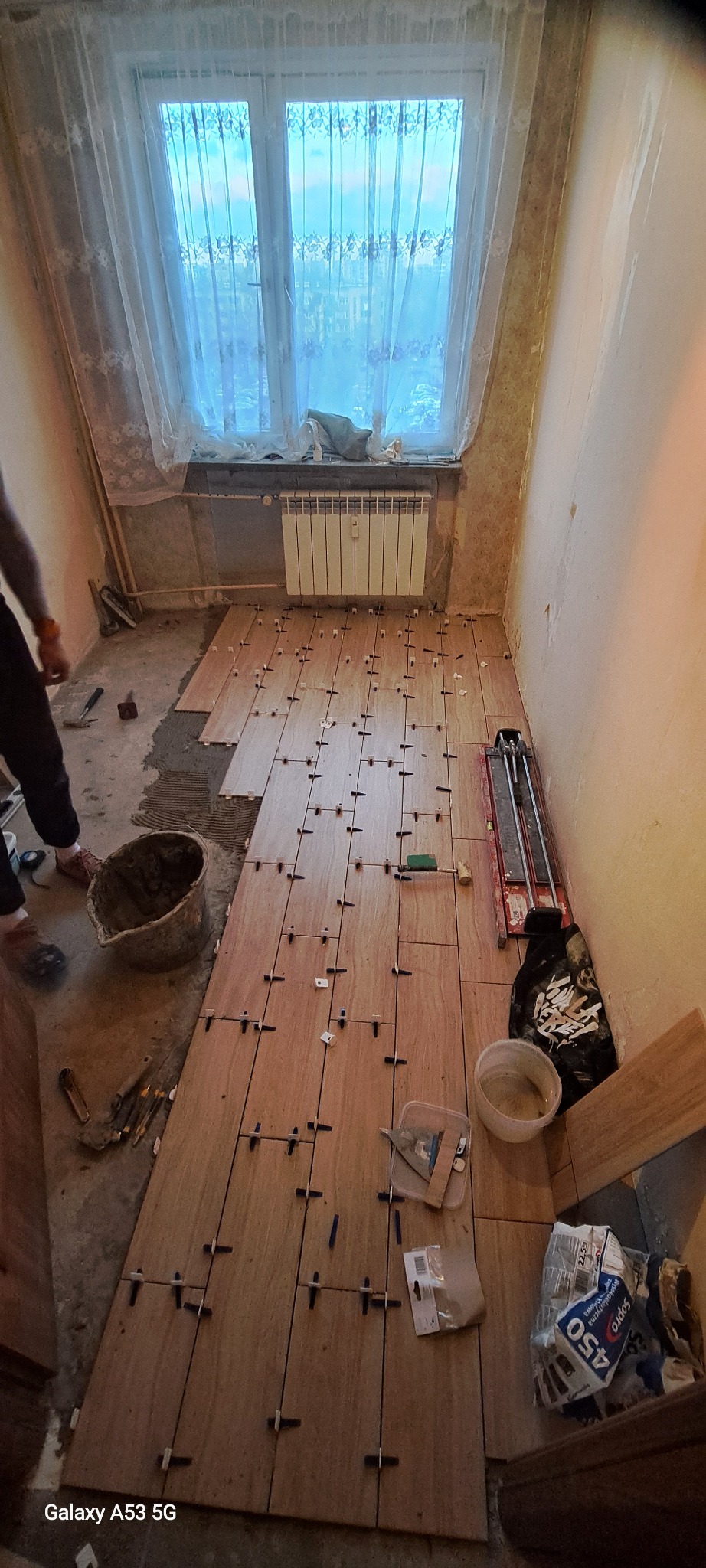 Układanie podłogi z płytek imitujących drewno w pokoju z oknem, widoczne krzyżyki dystansowe, wiadro z zaprawą, narzędzia glazurnicze oraz częściowo ułożone płytki.