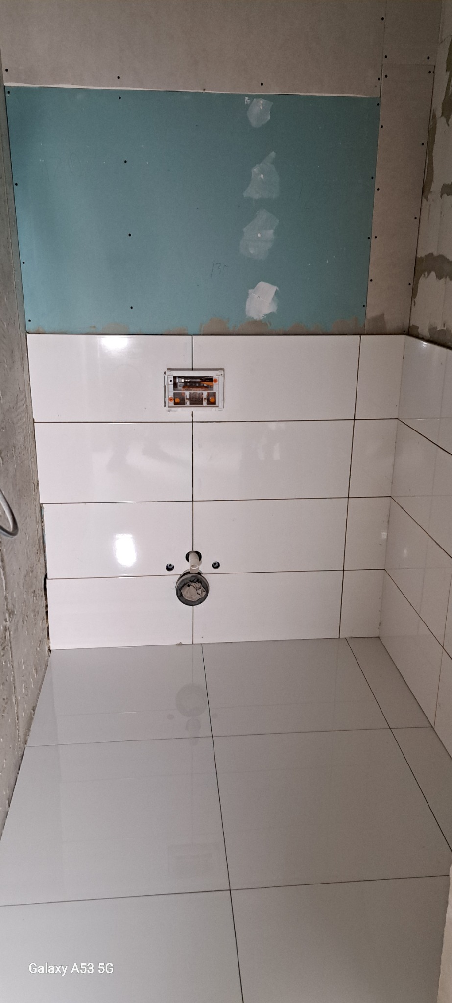 Białe płytki ścienne i podłogowe w łazience w trakcie wykańczania, widoczna instalacja podtynkowa i fragment ściany z płyty gipsowo-kartonowej z widocznymi śladami po szpachlowaniu.