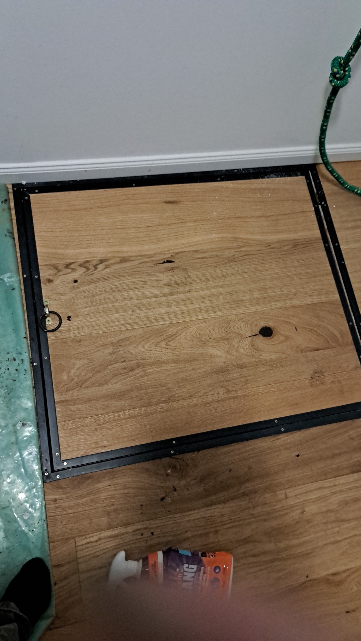 Drewniana klapa podłogowa z czarną metalową ramą i uchwytem pierścieniowym, widoczna na tle podłogi z paneli, z resztkami pyłu i pojemnikiem z pianką montażową w dolnej części kadru.