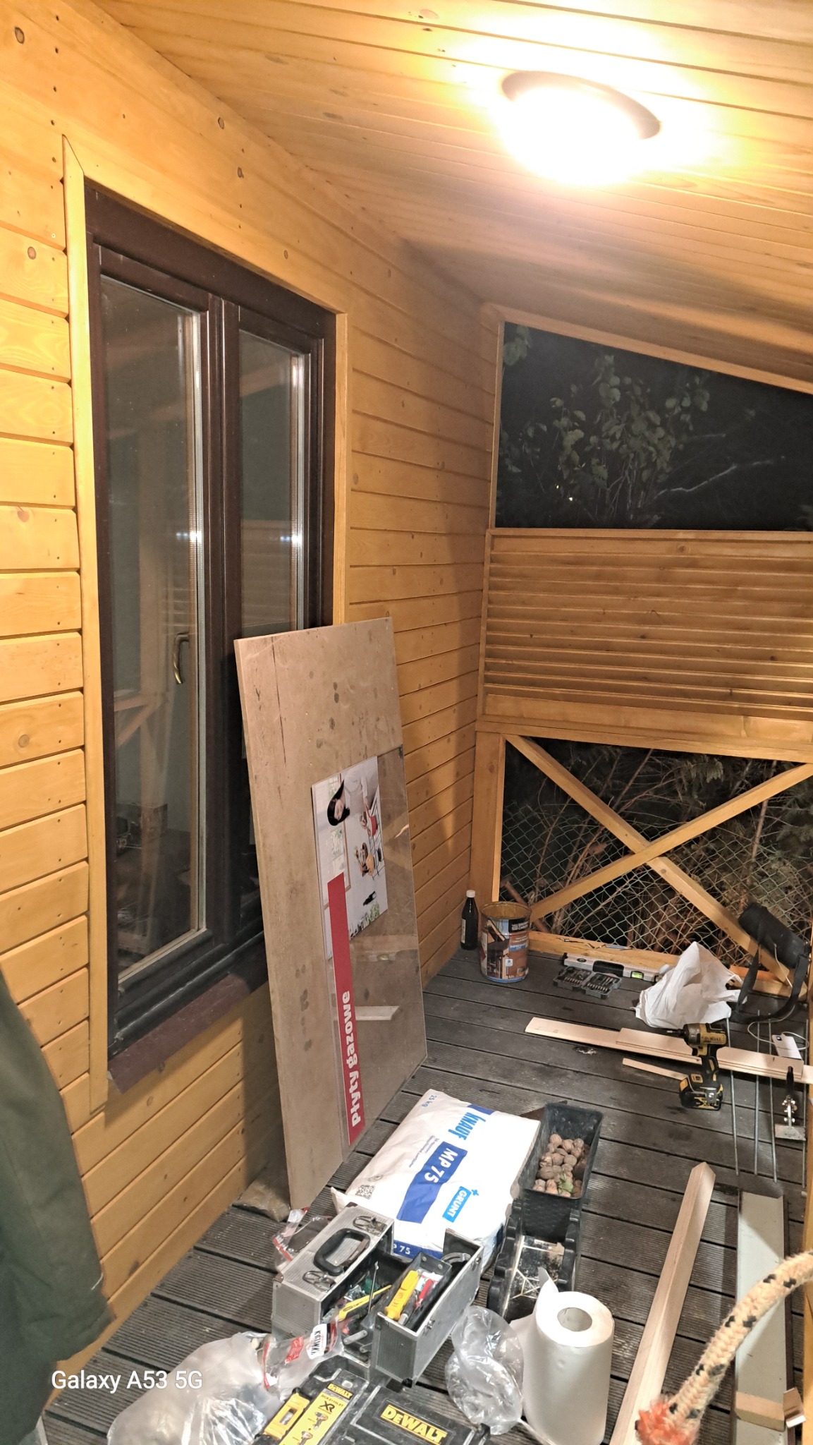 Wnętrze drewnianej konstrukcji w trakcie prac wykończeniowych: widoczne okno z brązową ramą, płyta gipsowa oparta o ścianę, narzędzia budowlane i materiały wykończeniowe, sufit i ściany...