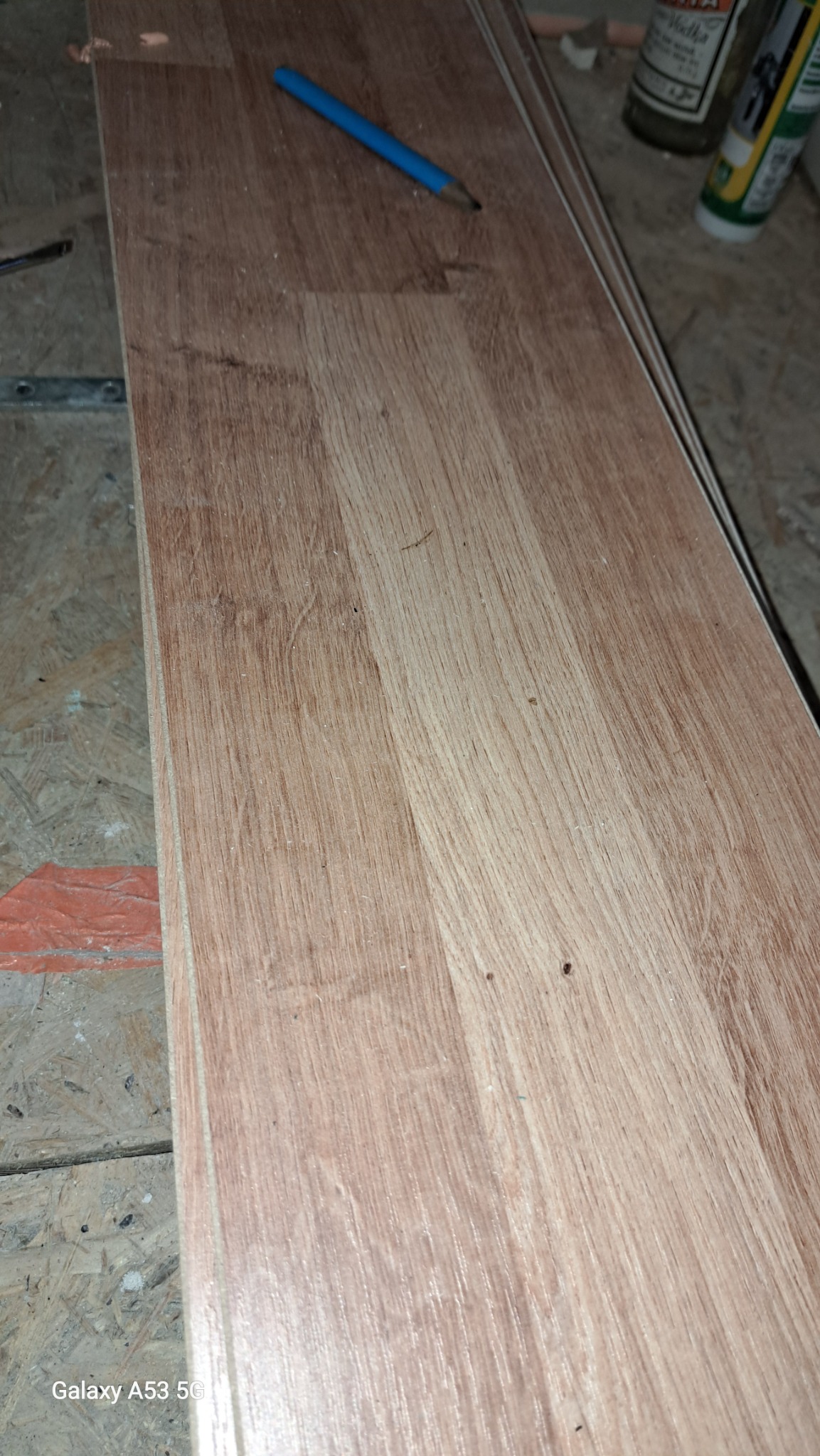 Panele podłogowe imitujące drewno, ułożone jeden na drugim, z niebieskim ołówkiem na wierzchu, widoczne butelki z klejem w tle.