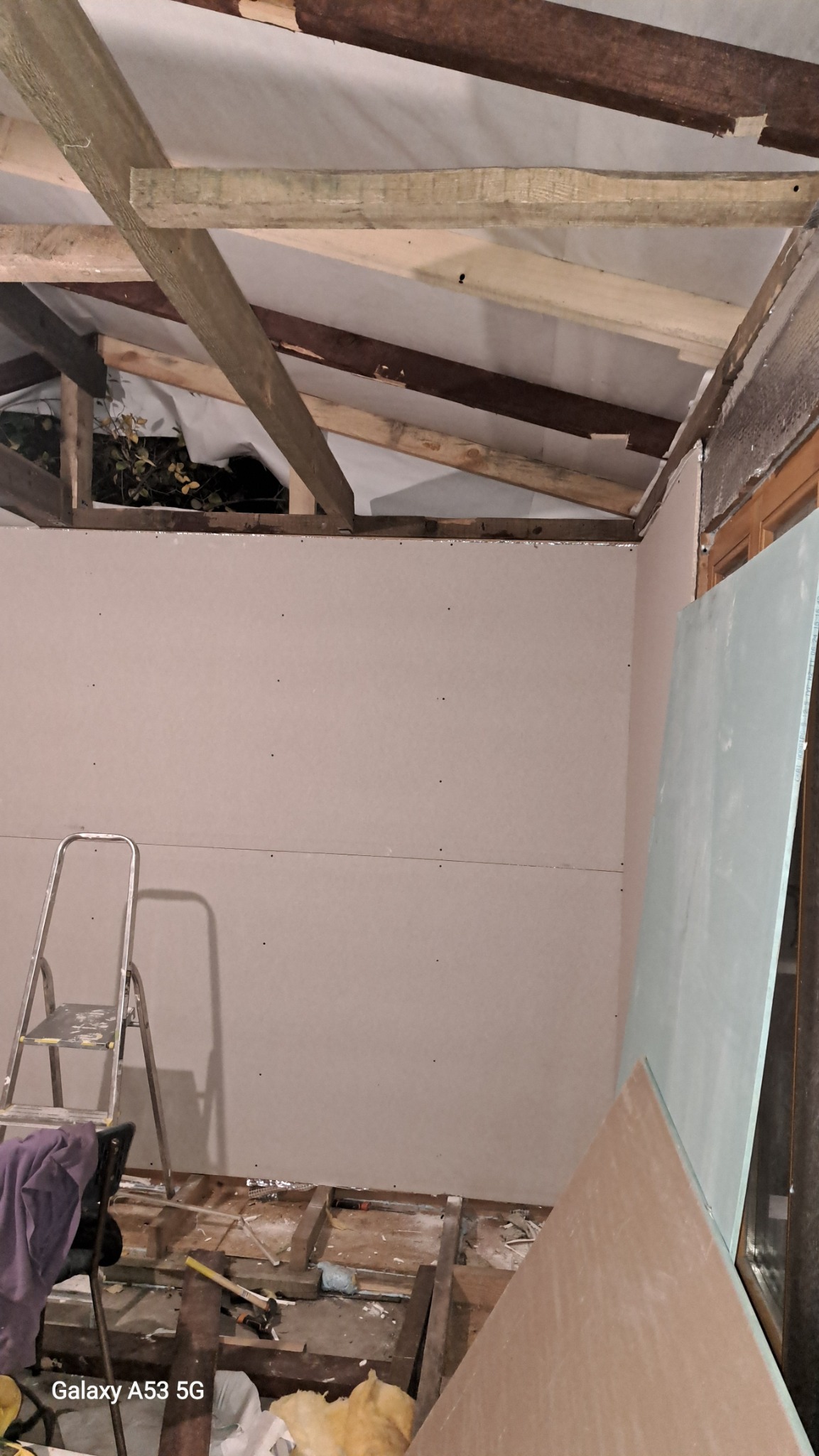 Wnętrze w trakcie prac wykończeniowych: ściany z płyt kartonowo-gipsowych, widoczne drewniane elementy konstrukcyjne dachu, drabina i bałagan na podłodze.