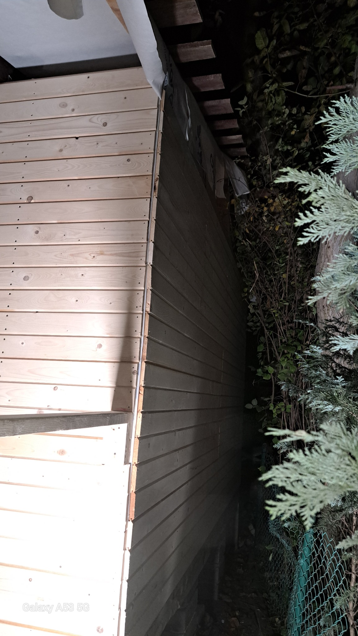 Fragment drewnianej elewacji budynku w trakcie montażu, widoczne deski boazeryjne, folia wiatroizolacyjna, ciemne otoczenie z roślinnością.