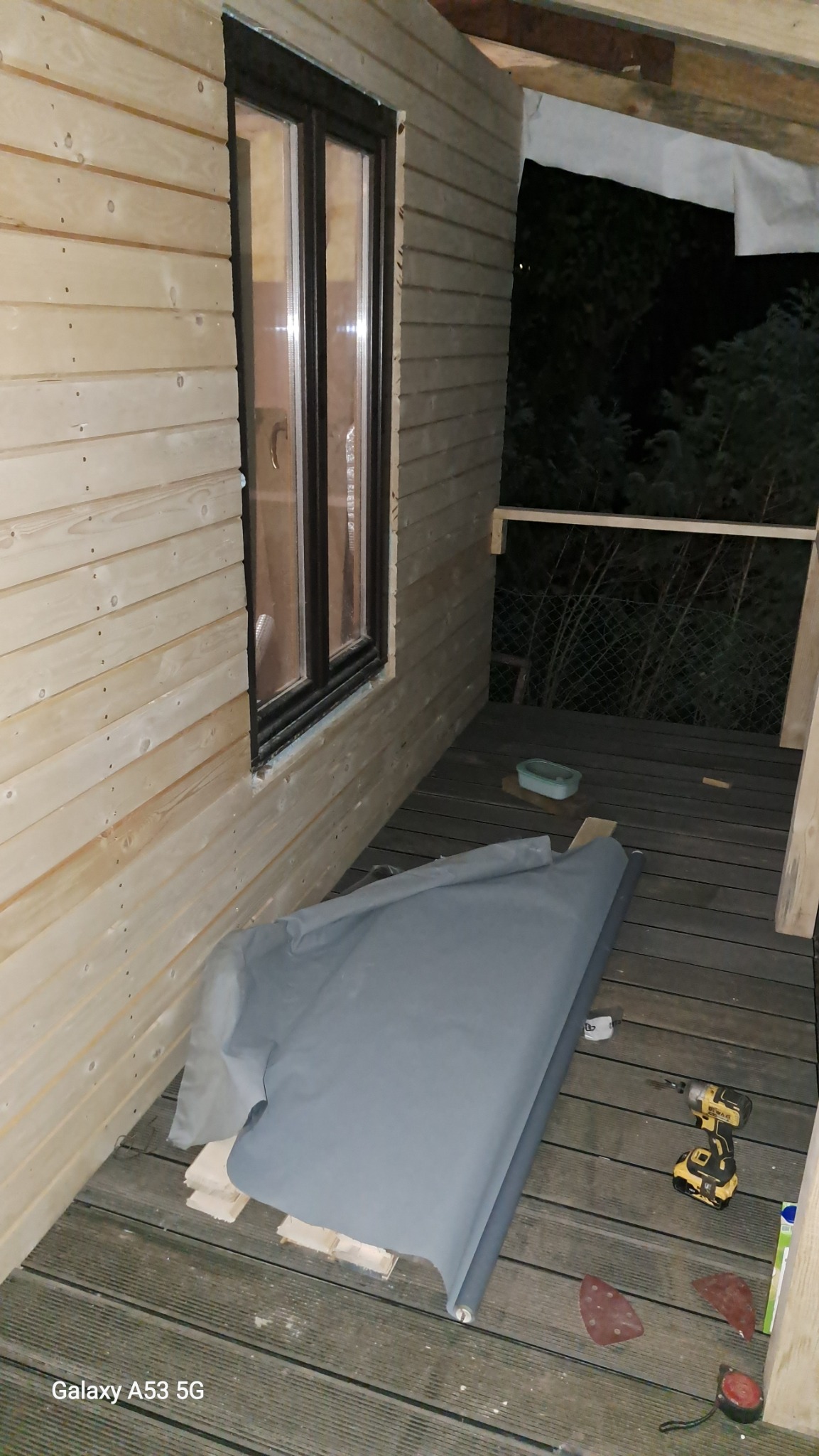 Drewniana elewacja budynku z zamontowanym oknem, częściowo zakryta szarą folią ochronną, w tle widoczne drzewa, na podłodze wiertarka i papier ścierny.