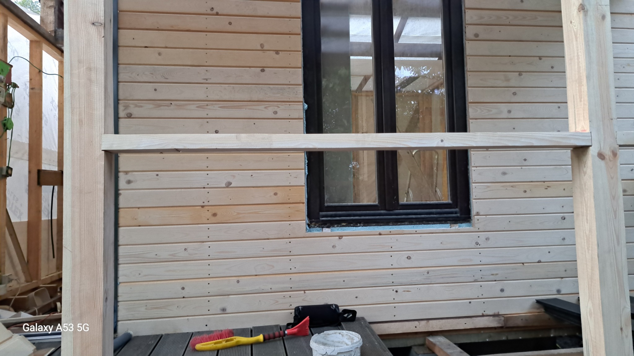 Drewniana elewacja domu w trakcie montażu, widoczne deski elewacyjne z jasnego drewna, czarna rama okienna z niezaizolowaną szczeliną, elementy konstrukcyjne z drewna, narzędzia pracy (szczotka...