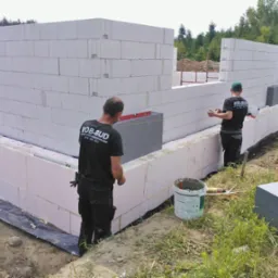 Budowa domu pasywnego: Dwóch robotników w czarnych koszulkach z logo firmy wznosi ściany z białych bloczków, izolowane szarym styropianem, widoczne narzędzia i wiadro z zaprawą.