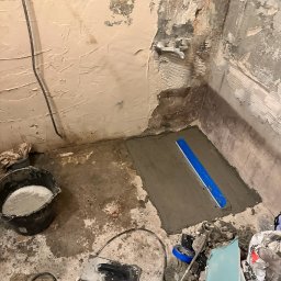 Jakub krasowski - Przygotowanie podłoża pod prysznic w trakcie prac, widoczna wylewka wyrównana łatą, surowe ściany, instalacja wodna, kubeł z zaprawą, narzędzia i worki na gruz.