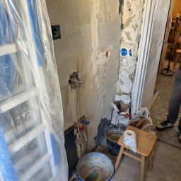 Jakub krasowski - Niedokończona ściana w trakcie przygotowania do prac wykończeniowych, widoczne odsłonięte rury, drabina zabezpieczona folią, wiadro z zaprawą, rolka papieru toaletowego na taborecie i resztki...
