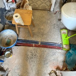 Jakub krasowski - Łazienka w trakcie remontu: widoczne rury, wiadro z zaprawą, mop, płytki, rolka papieru toaletowego na taborecie i stopa w roboczym bucie.