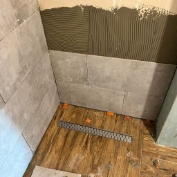 Jakub krasowski - Układanie szarych płytek w kabinie prysznicowej, widoczny odpływ liniowy, świeżo nałożony klej na ścianie i krzyżyki dystansowe.