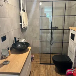 Nowoczesna łazienka z szarą płytką, czarną umywalką w stylu marmuru, kabiną prysznicową z czarnymi ramami i drewnopodobną podłogą.