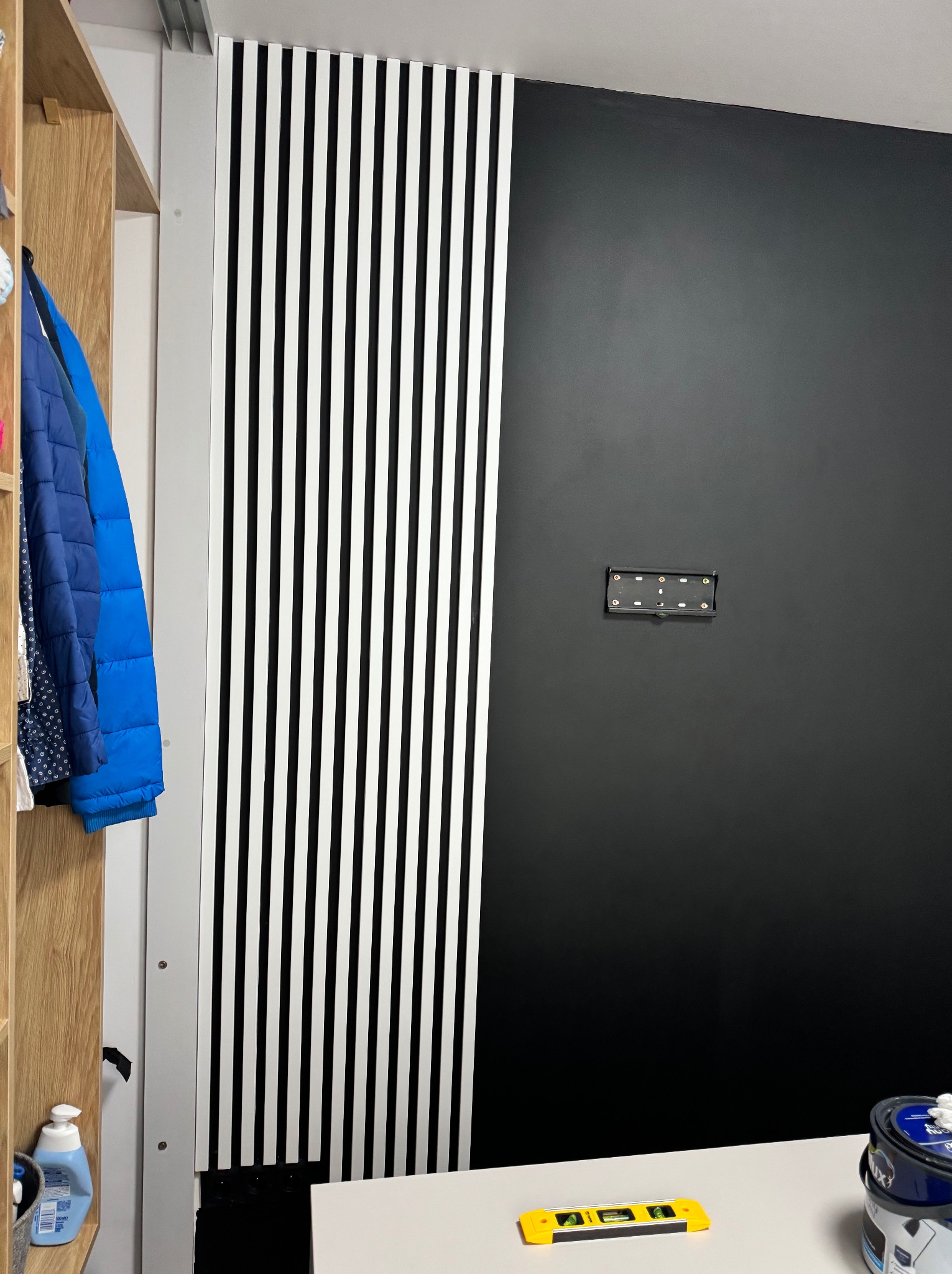 Częściowo wykończona ściana z czarną farbą i dekoracyjnymi białymi listwami, widoczny uchwyt do telewizora, poziomnica i puszka z farbą na blacie.