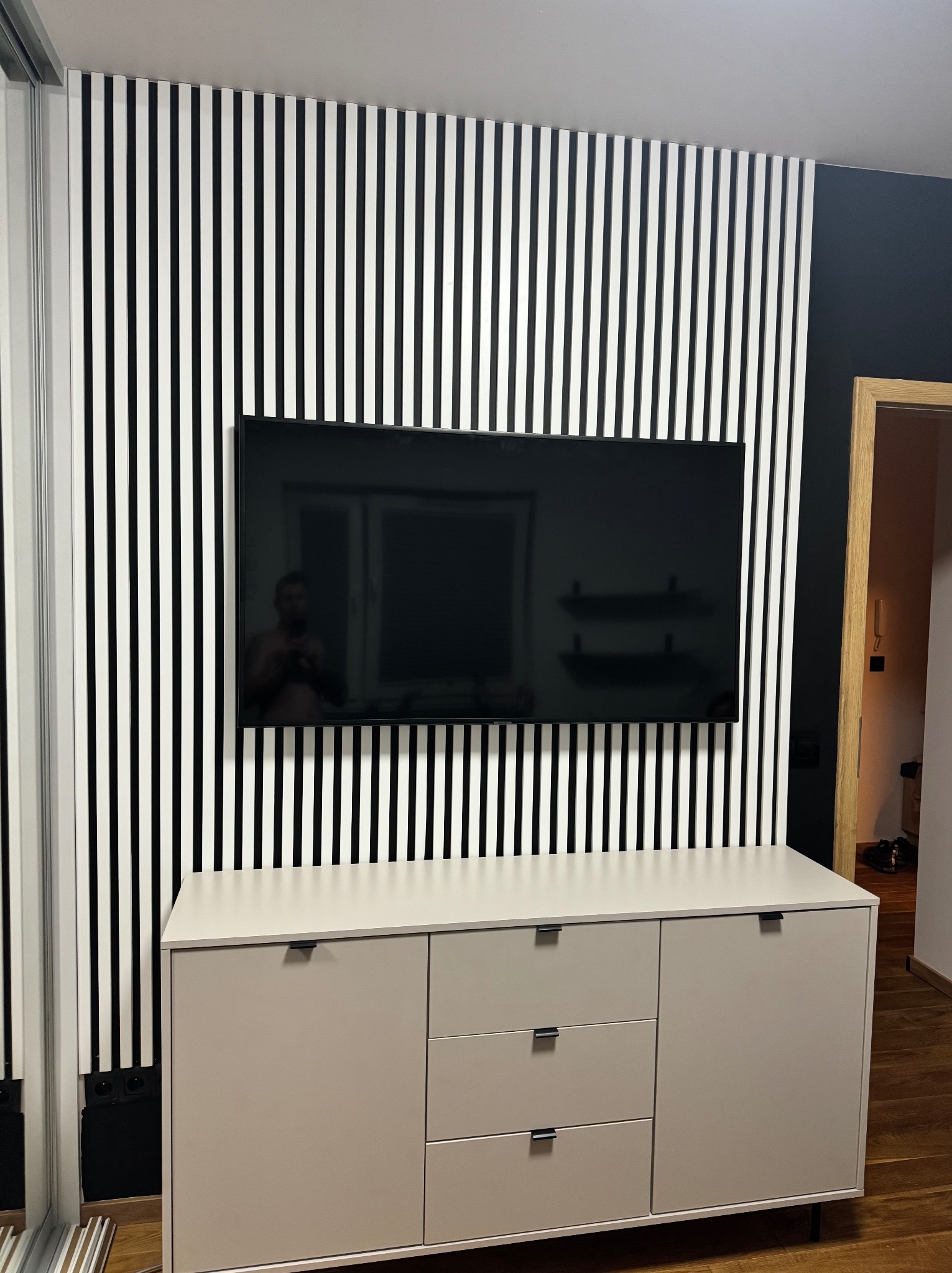 Nowoczesny salon z dekoracyjną ścianą z białych listew na czarnym tle, na której wisi telewizor, pod telewizorem stoi beżowa komoda z trzema szufladami i szafką, widoczna podłoga z paneli...