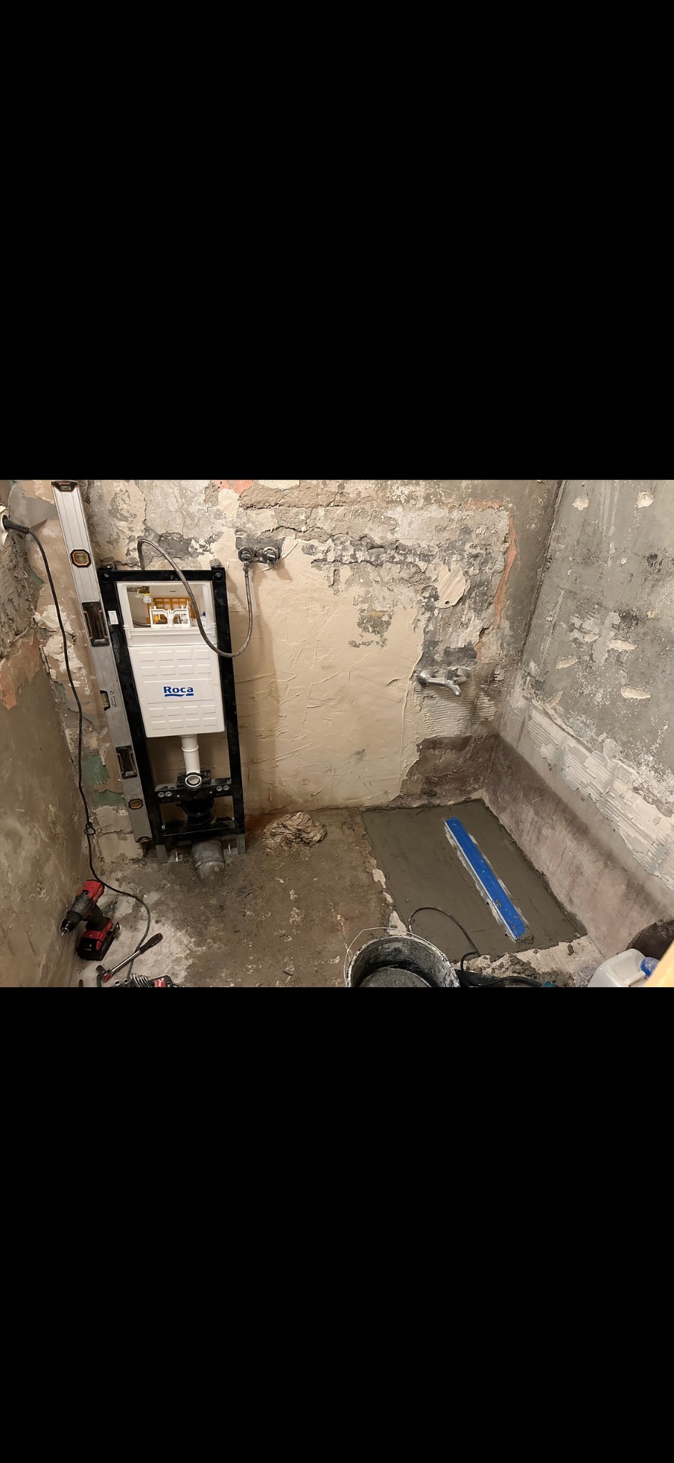 Nierówna ściana łazienki w trakcie renowacji z zamontowanym stelażem podtynkowym Roca i wylewką pod prysznic, widoczne narzędzia i poziomica.