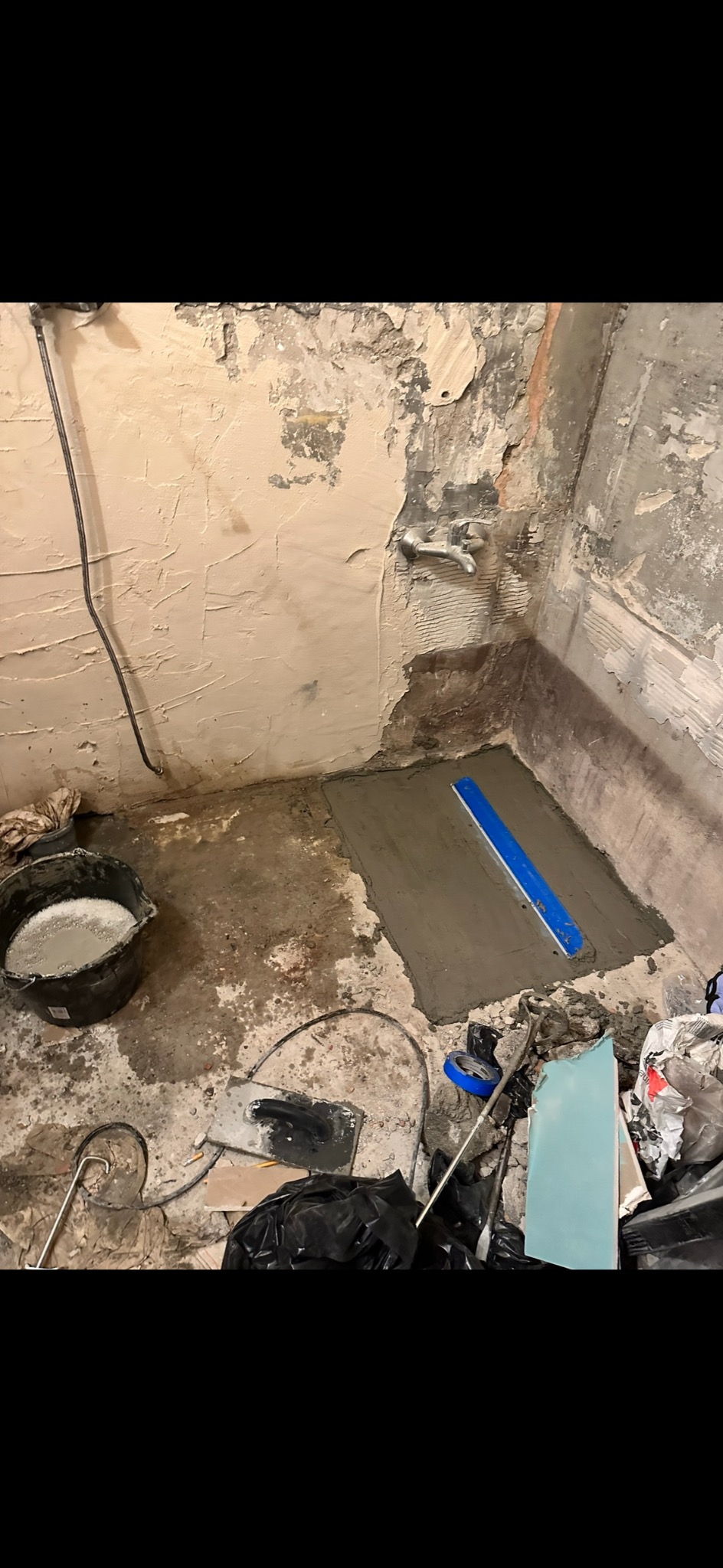 Przygotowanie podłoża pod prysznic w trakcie prac, widoczna wylewka wyrównana łatą, surowe ściany, instalacja wodna, kubeł z zaprawą, narzędzia i worki na gruz.