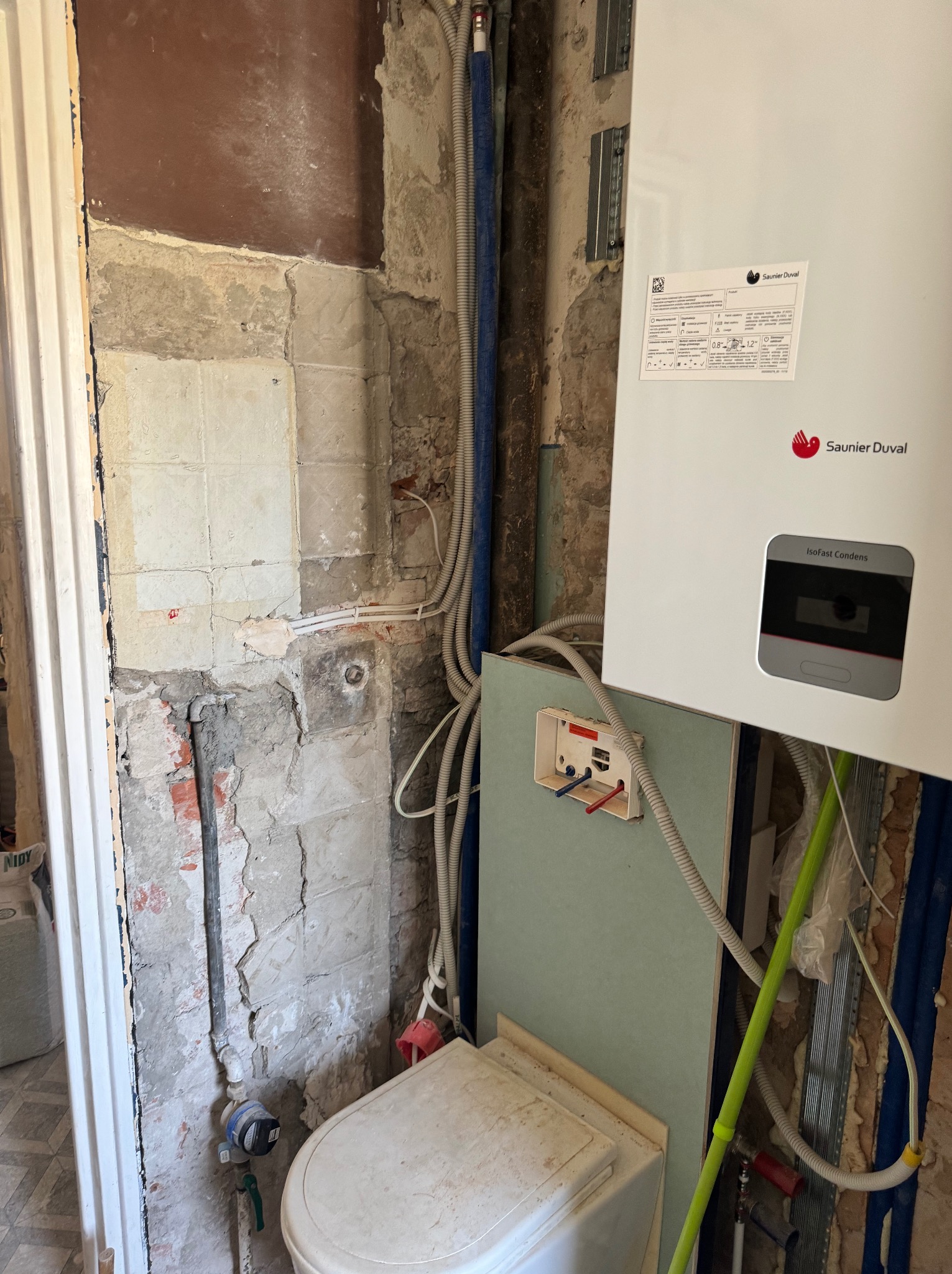 Wnętrze łazienki w trakcie montażu nowego pieca gazowego Saunier Duval, widoczne odsłonięte ściany z fragmentami starych płytek, rury instalacyjne i częściowo zamontowana toaleta.
