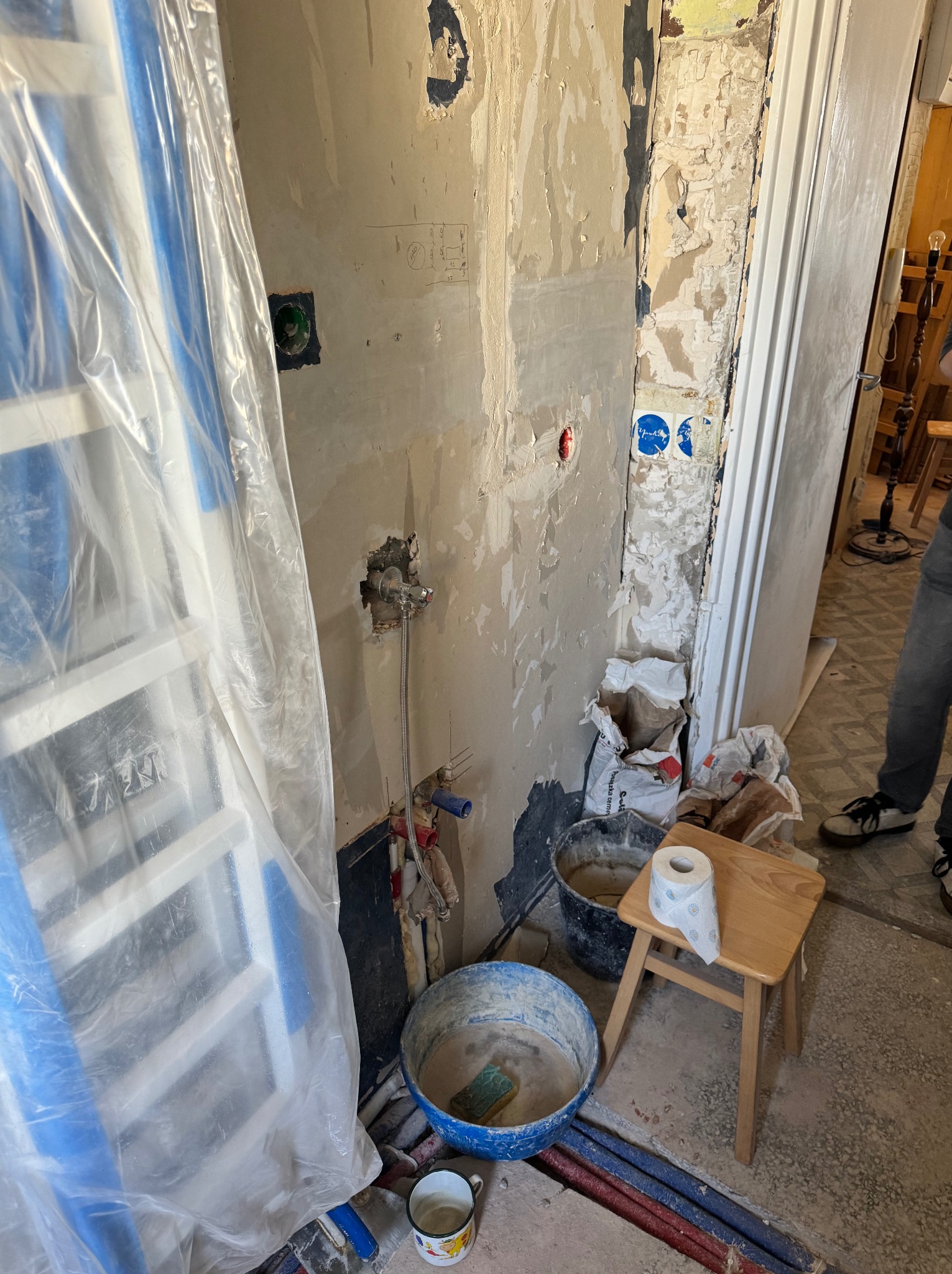 Niedokończona ściana w trakcie przygotowania do prac wykończeniowych, widoczne odsłonięte rury, drabina zabezpieczona folią, wiadro z zaprawą, rolka papieru toaletowego na taborecie i resztki...