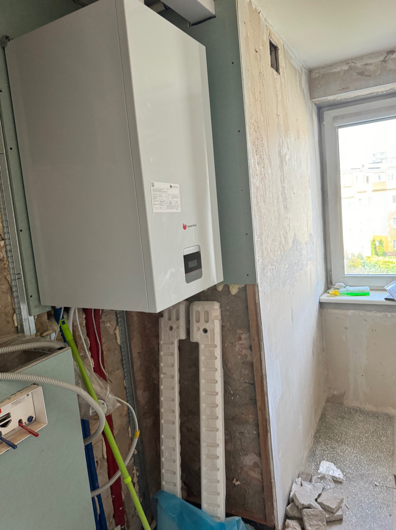 Montaż nowego pieca gazowego marki Saunier Duval na ścianie z płyt gipsowo-kartonowych, widoczne rury instalacyjne w kolorach czerwonym, niebieskim i zielonym, w tle okno i surowe ściany w trakcie...