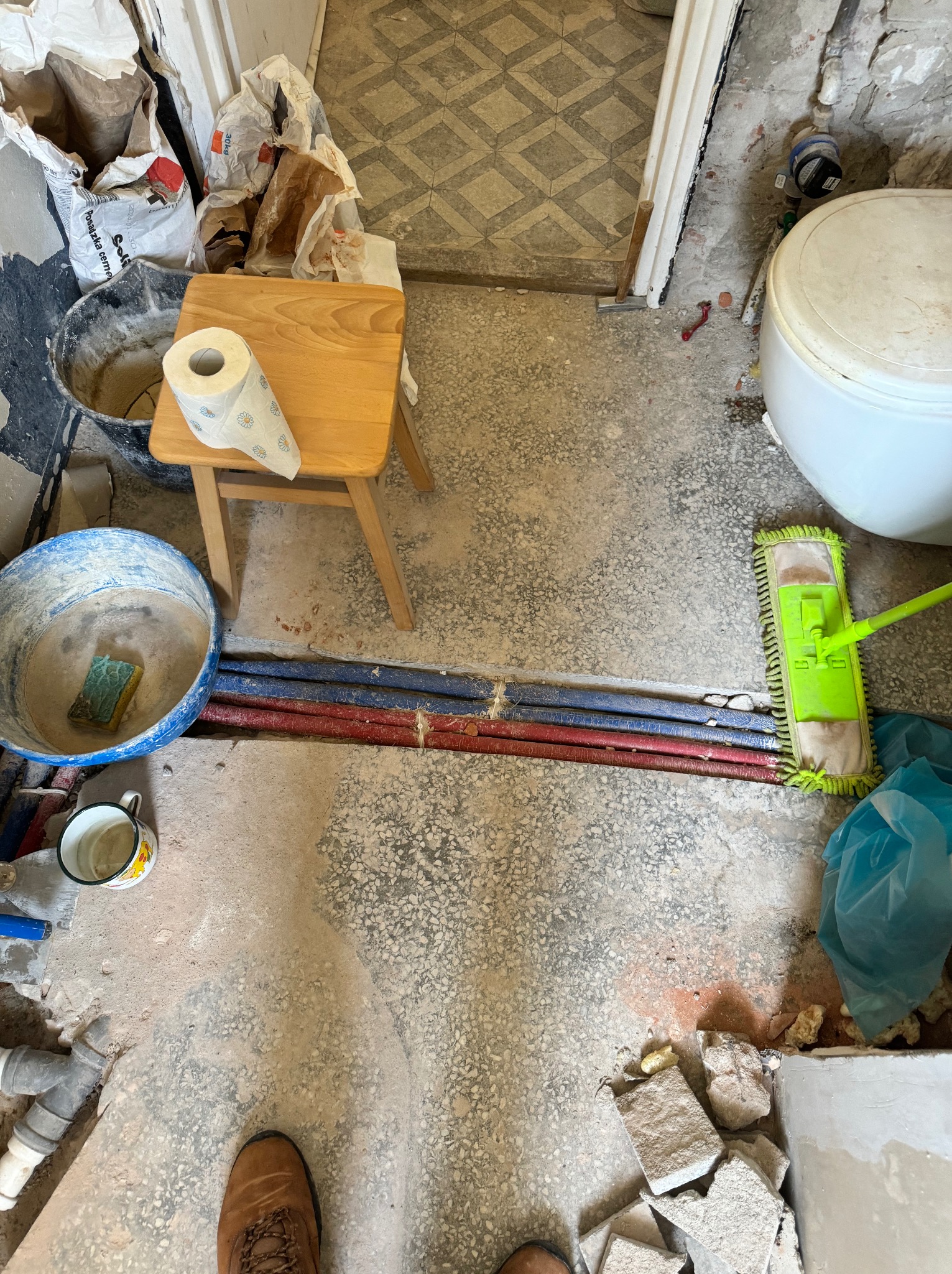 Łazienka w trakcie remontu: widoczne rury, wiadro z zaprawą, mop, płytki, rolka papieru toaletowego na taborecie i stopa w roboczym bucie.
