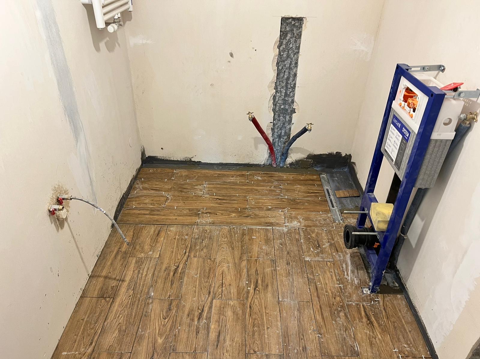 Łazienka w trakcie prac: ułożona podłoga z płytek imitujących drewno, widoczny stelaż podtynkowy WC, instalacja wodna w ścianie, grzejnik na ścianie.