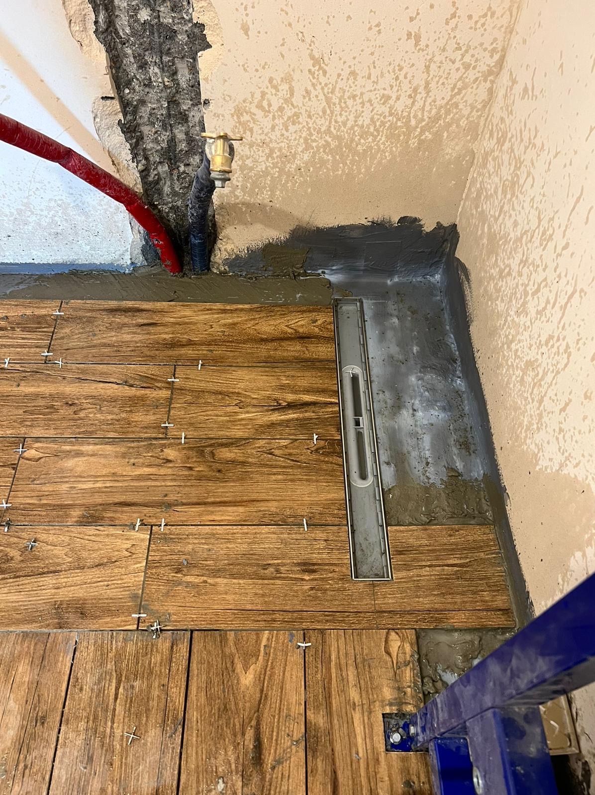 Ułożone drewnopodobne płytki podłogowe w łazience z liniowym odpływem, widoczne krzyżyki dystansowe, instalacja wodna w ścianie i fragment stelaża podtynkowego WC.