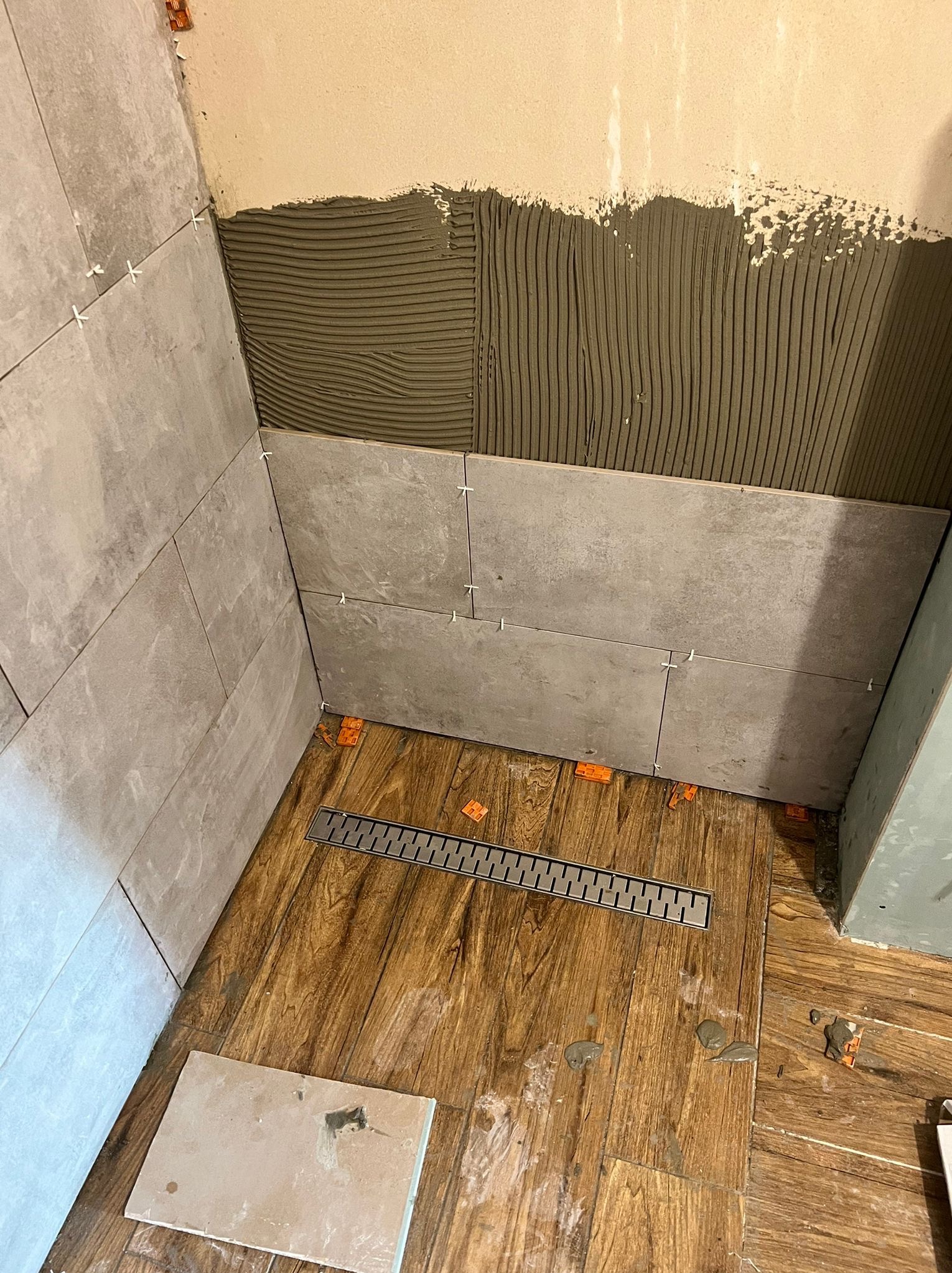 Układanie szarych płytek w kabinie prysznicowej, widoczny odpływ liniowy, świeżo nałożony klej na ścianie i krzyżyki dystansowe.