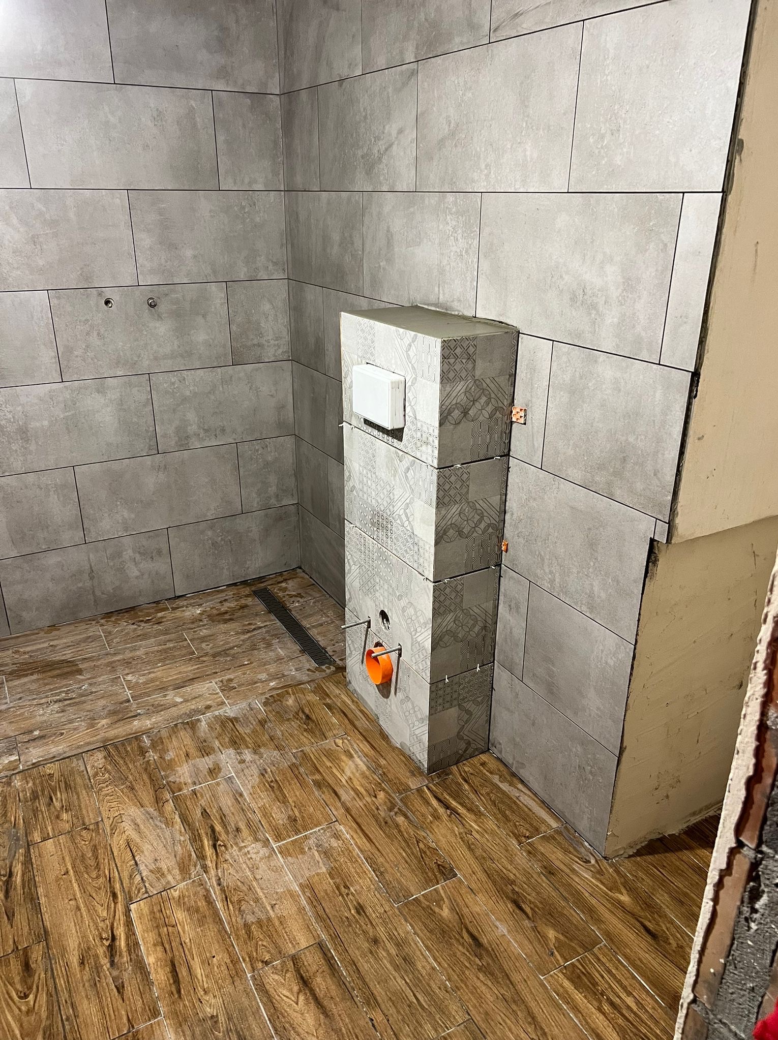 Ściana wyłożona szarymi płytkami w trakcie montażu, widoczne krzyżyki dystansowe, podłoga z płytek imitujących drewno, zabudowa stelaża WC z dekoracyjnymi płytkami.