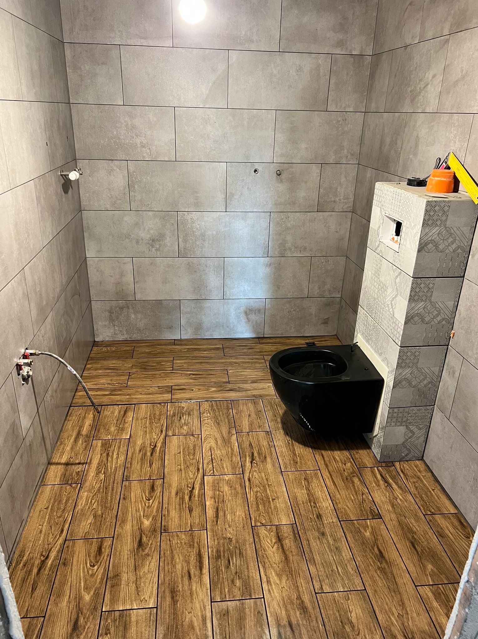 Nowoczesna łazienka w trakcie wykańczania: szare, prostokątne płytki ścienne, podłoga wyłożona panelami imitującymi drewno, czarna, podwieszana toaleta z zabudową w płytkach o wzorzystym designie...