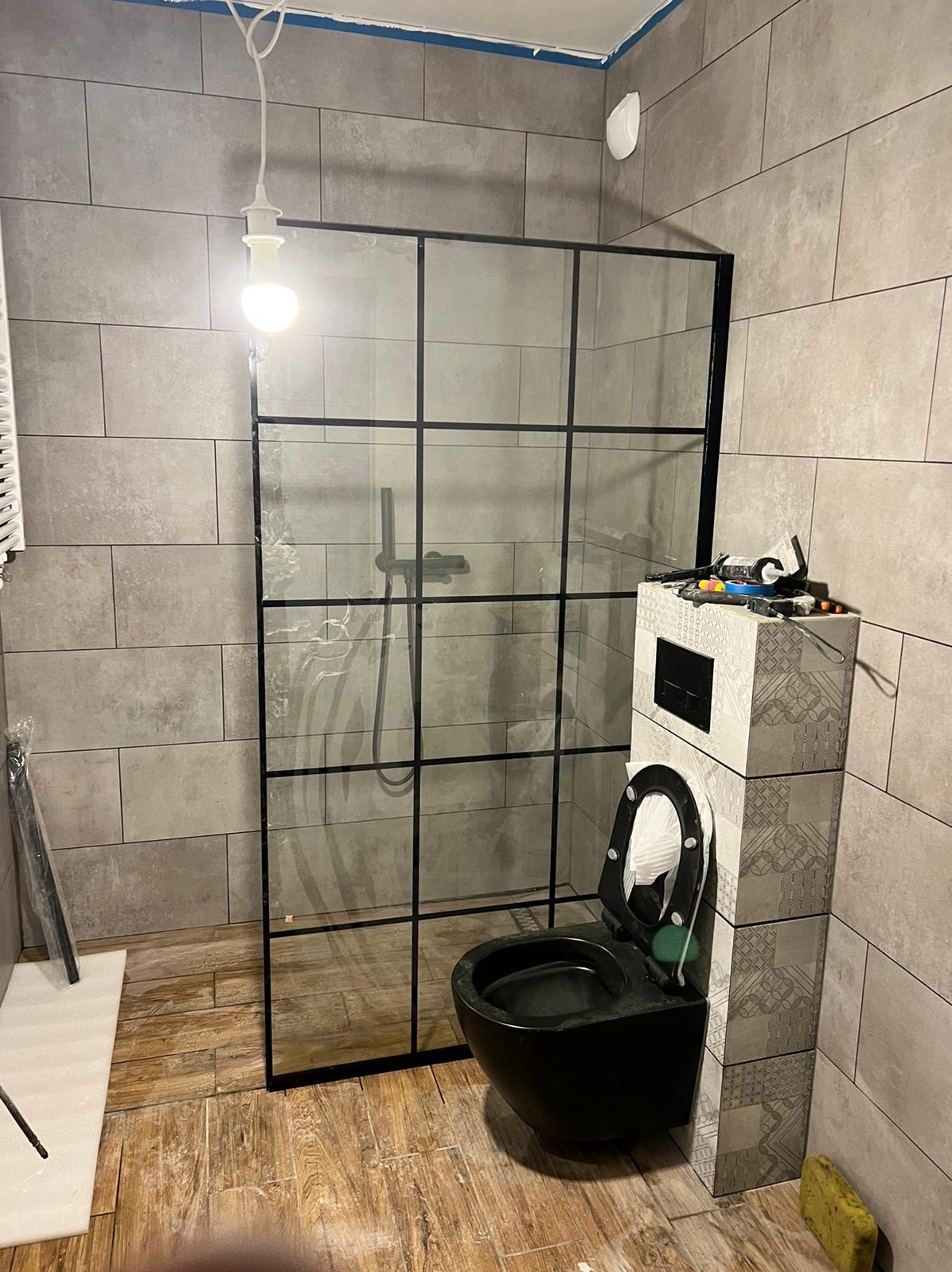Remont łazienki: Nowoczesna kabina prysznicowa z czarnymi ramami i czarna wisząca toaleta w trakcie montażu, widoczne narzędzia na spłuczce, szare płytki na ścianach i podłodze imitujące drewno.