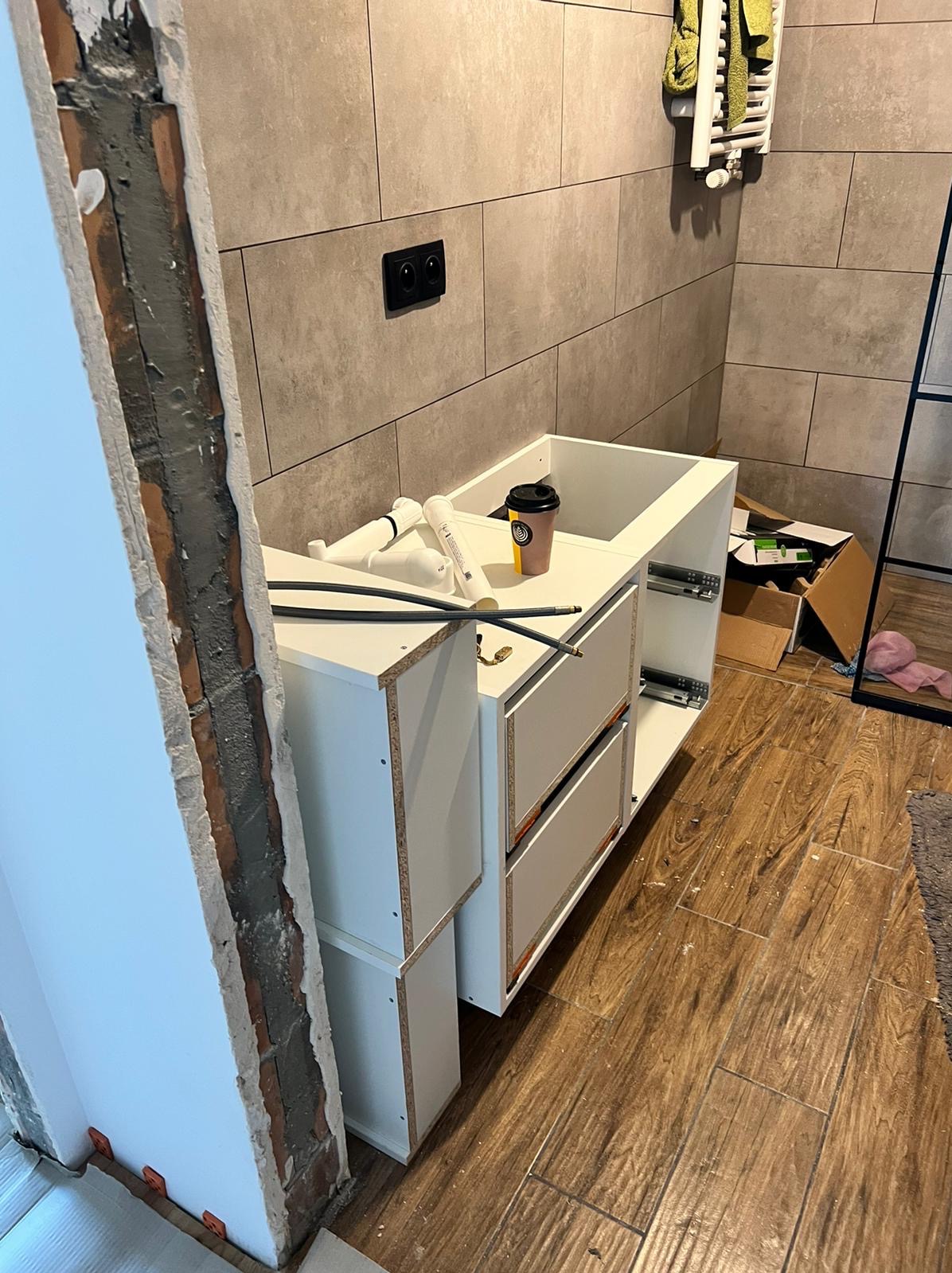 Montaż szafki pod umywalkę w łazience, widoczne niezakończone prace i odsłonięta ściana, na blacie kubek z kawą.