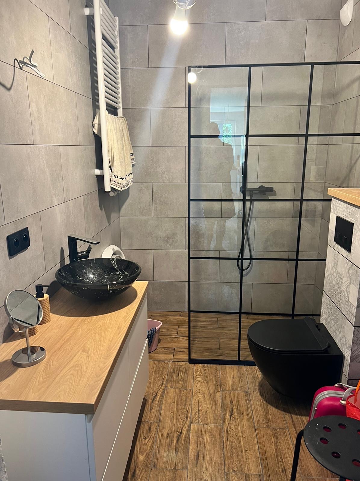 Nowoczesna łazienka z szarą płytką, czarną umywalką w stylu marmuru, kabiną prysznicową z czarnymi ramami i drewnopodobną podłogą.