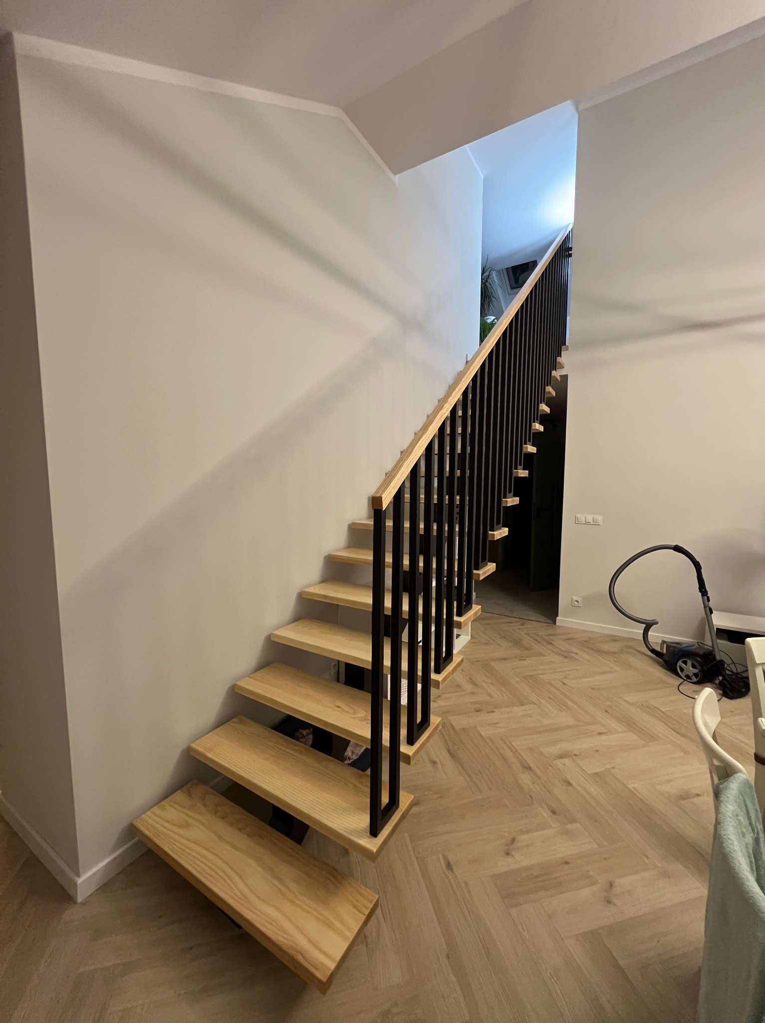 Nowoczesne schody drewniane z czarną, metalową konstrukcją, widoczne z boku, z parkietem w jodełkę na podłodze i odkurzaczem w tle.