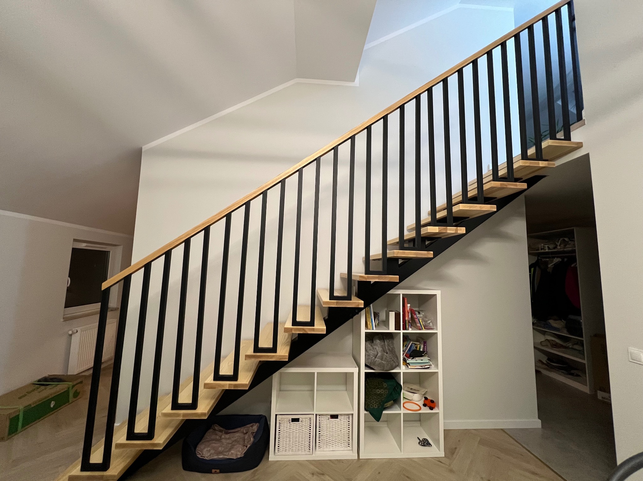 Nowoczesne schody z drewnianymi stopniami i czarną, minimalistyczną konstrukcją, widoczne z dołu, z elementami wystroju wnętrza w tle.