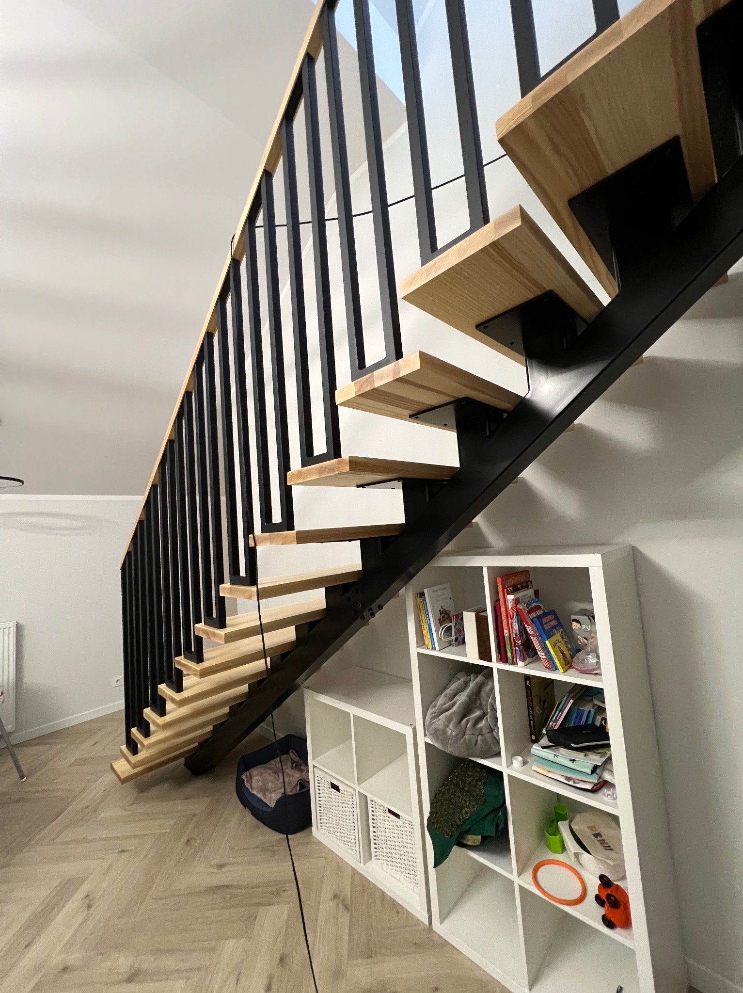 Nowoczesne schody wewnętrzne z drewnianymi stopniami i czarną, metalową konstrukcją, widoczne z dołu, z elementami wyposażenia wnętrza w tle.