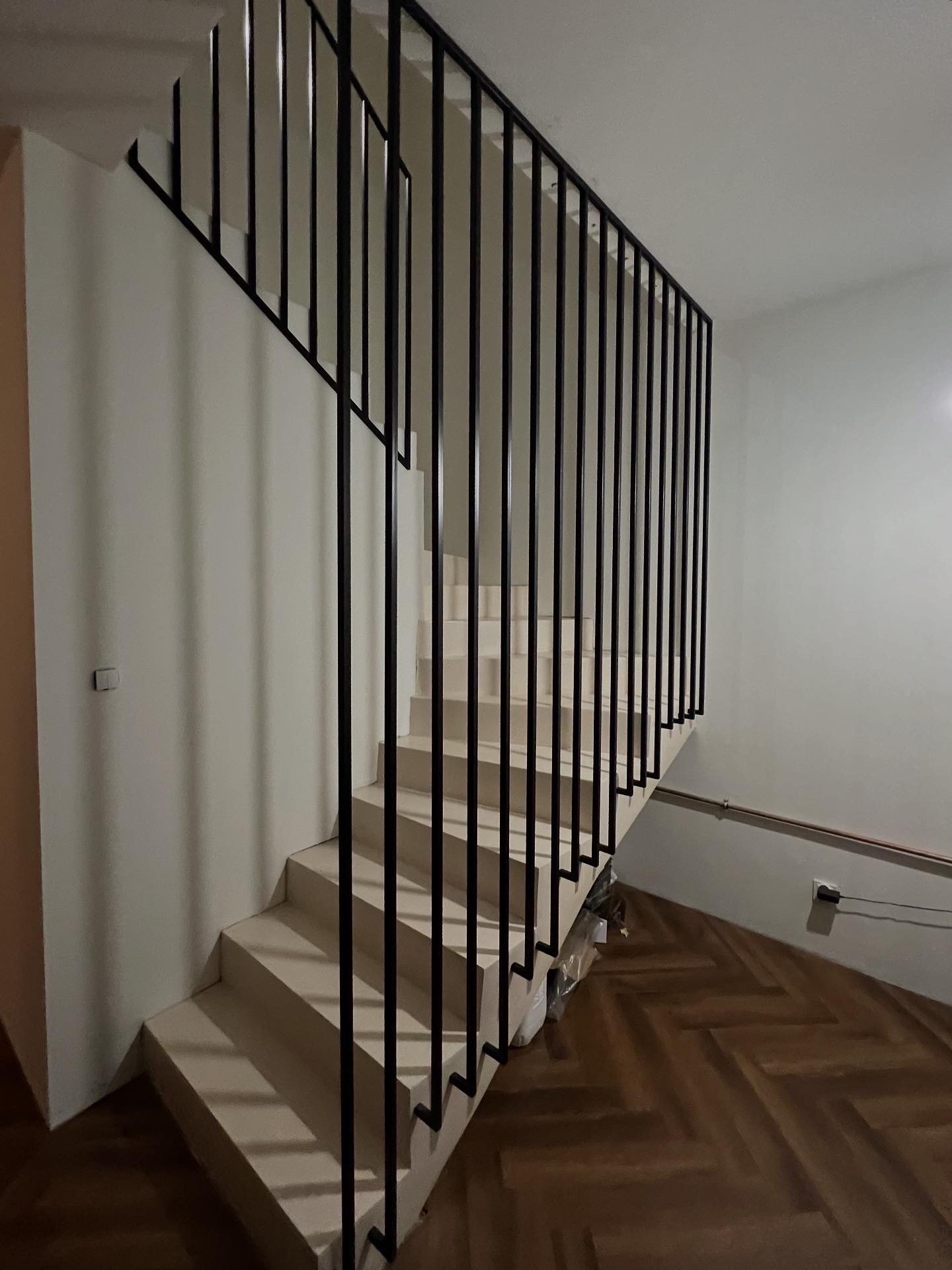 Minimalistyczne schody z czarną, pionową konstrukcją zabezpieczającą. Jasne stopnie kontrastują z ciemnym drewnem parkietu i białymi ścianami. Nowoczesny design.