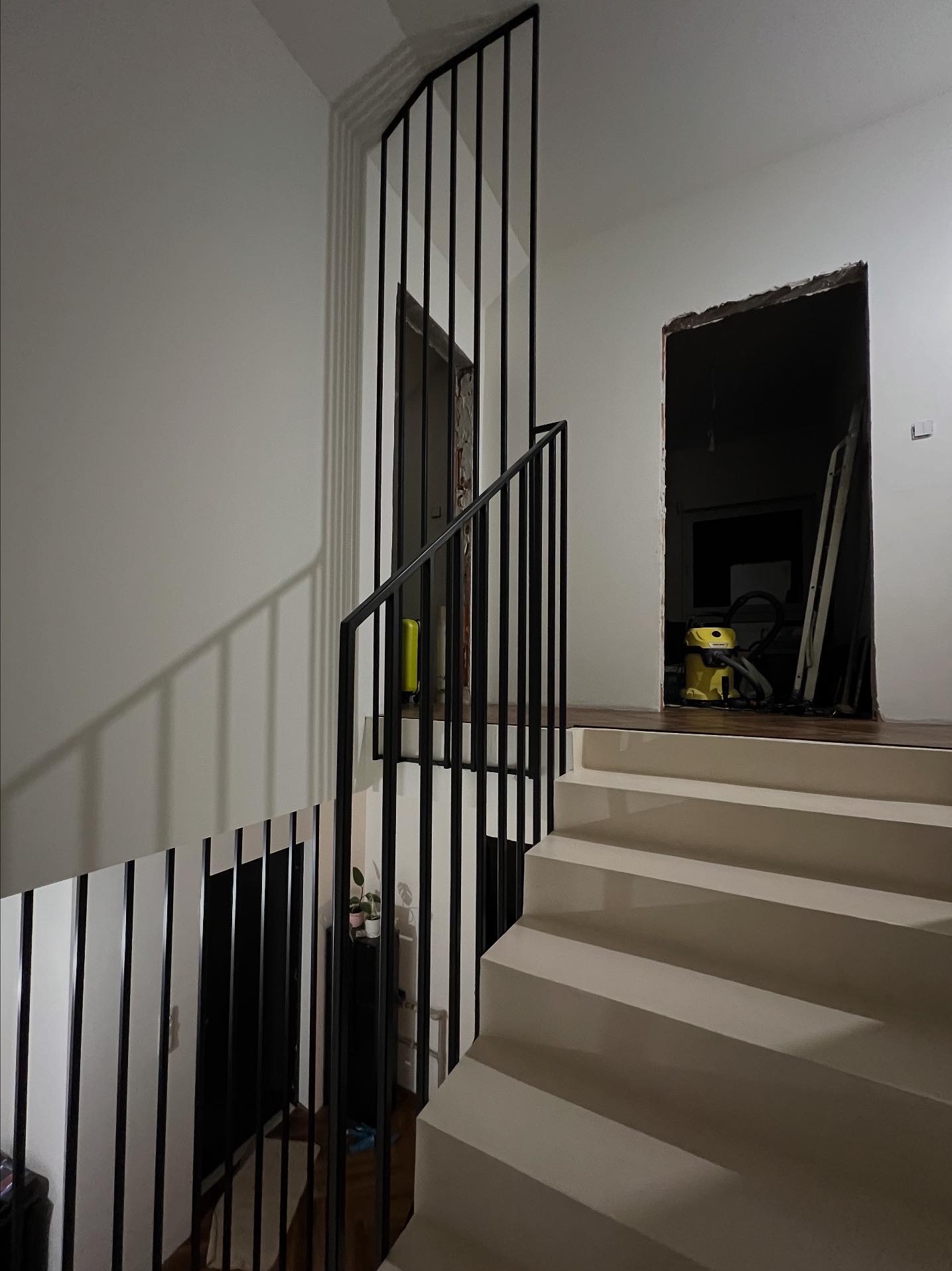 Minimalistyczne schody z czarną, pionową konstrukcją. Widoczne schody w jasnym odcieniu, fragment ściany i drzwi w tle. Ujęcie z dołu, podkreślające geometrię.