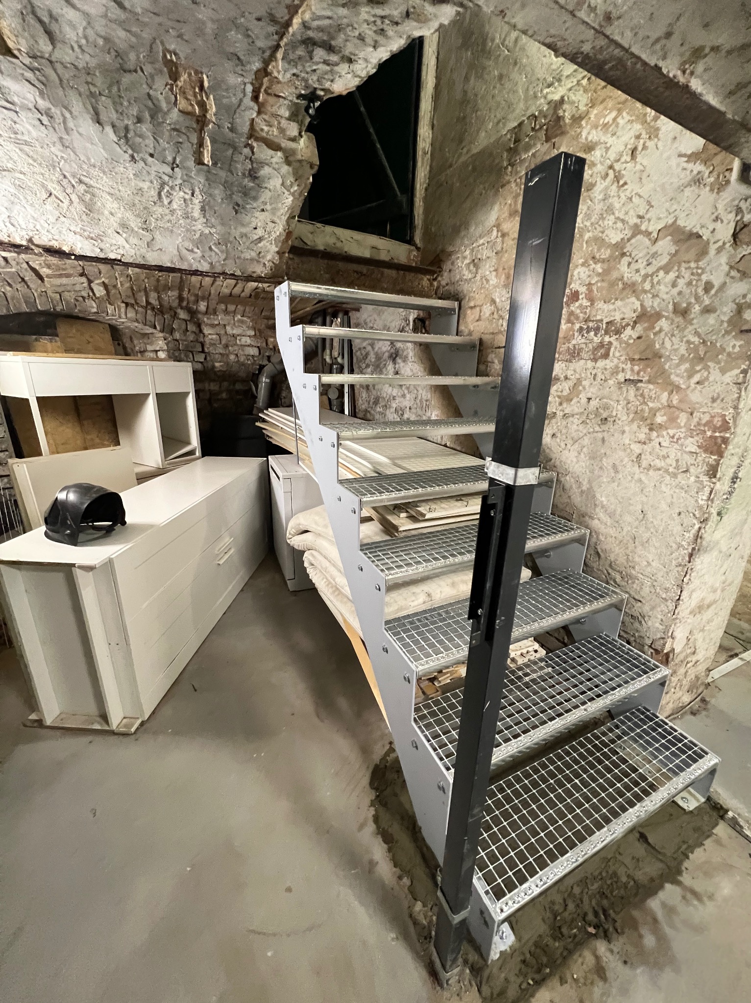 Metalowe schody kratowe w surowym wnętrzu z ceglanymi ścianami i elementami wyposażenia. Widok z dołu, ukazujący konstrukcję i otoczenie.