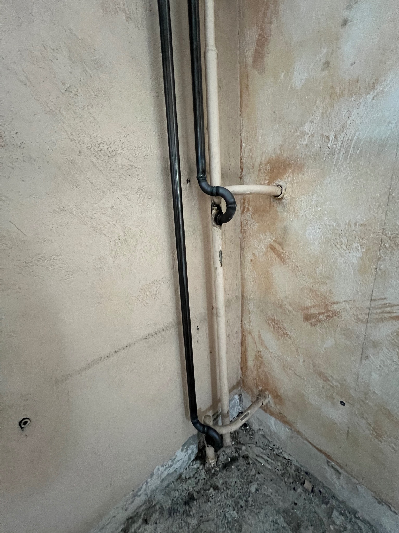 Narożne ułożenie rur w surowym wnętrzu. Dwie pionowe rury: czarna i beżowa, połączone czarnymi kolankami. Ściany z widocznymi śladami po pracach wykończeniowych.