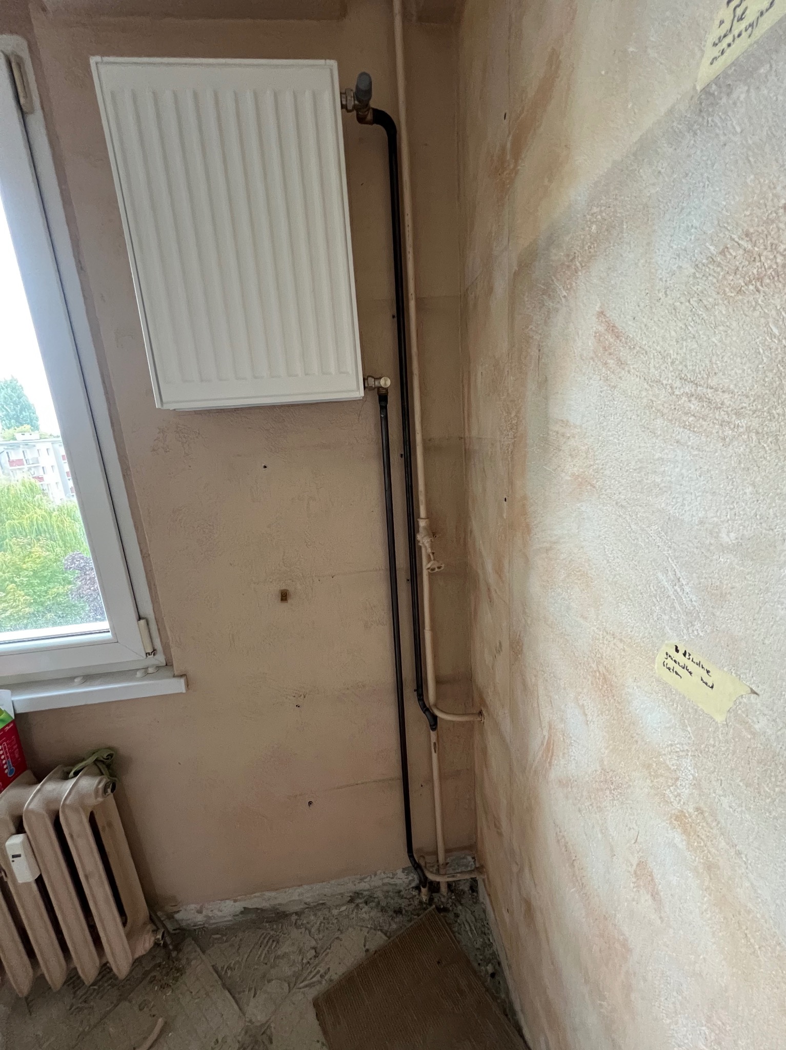 Biały grzejnik panelowy zamontowany na ścianie, z widocznymi rurami doprowadzającymi. Stary grzejnik żeliwny w rogu. Ściana w trakcie prac wykończeniowych.