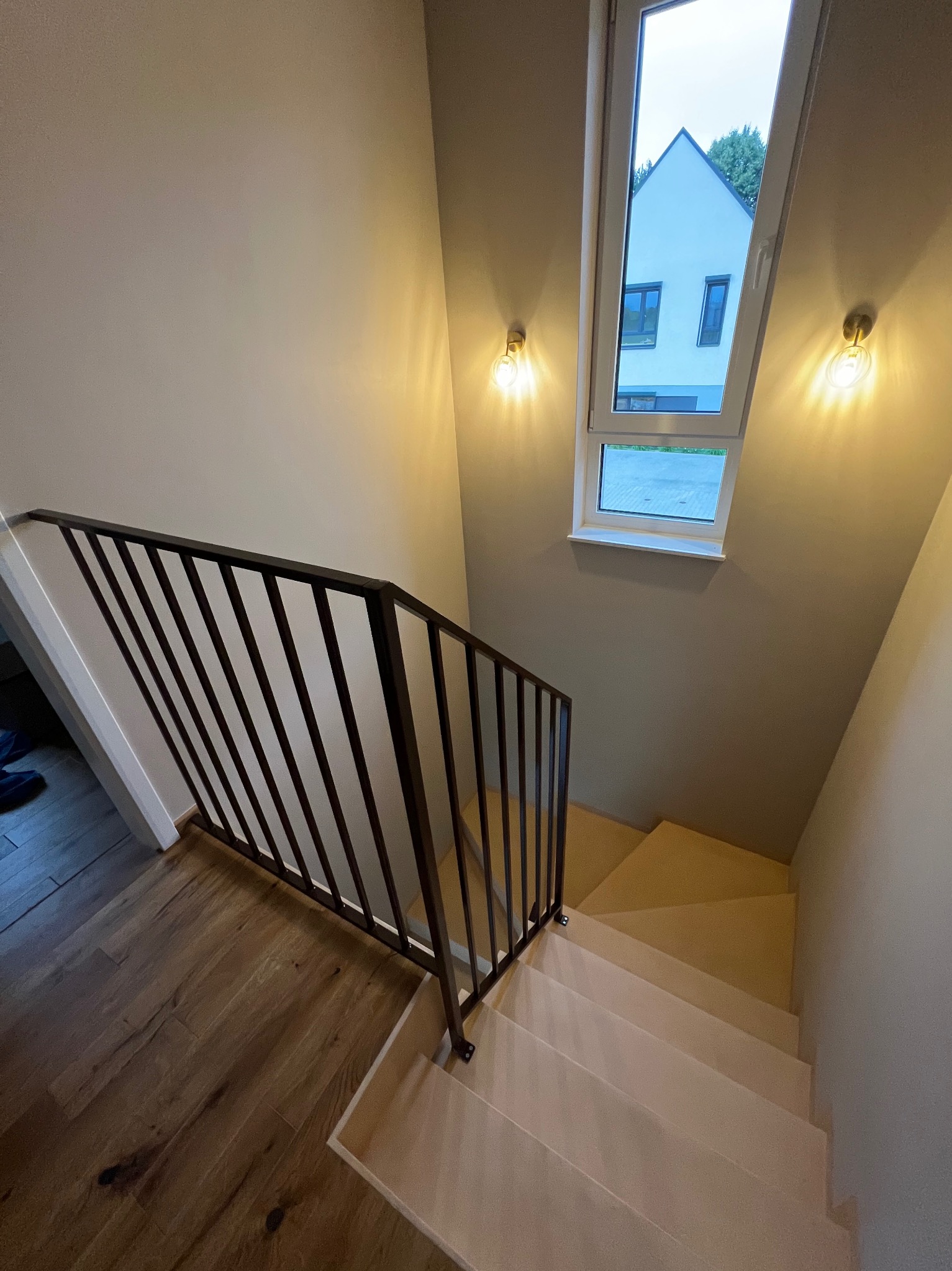 Nowoczesne schody z ciemną, metalową barierką i drewnianymi stopniami, oświetlone lampami ściennymi. Widok z góry na dół, z oknem w tle.