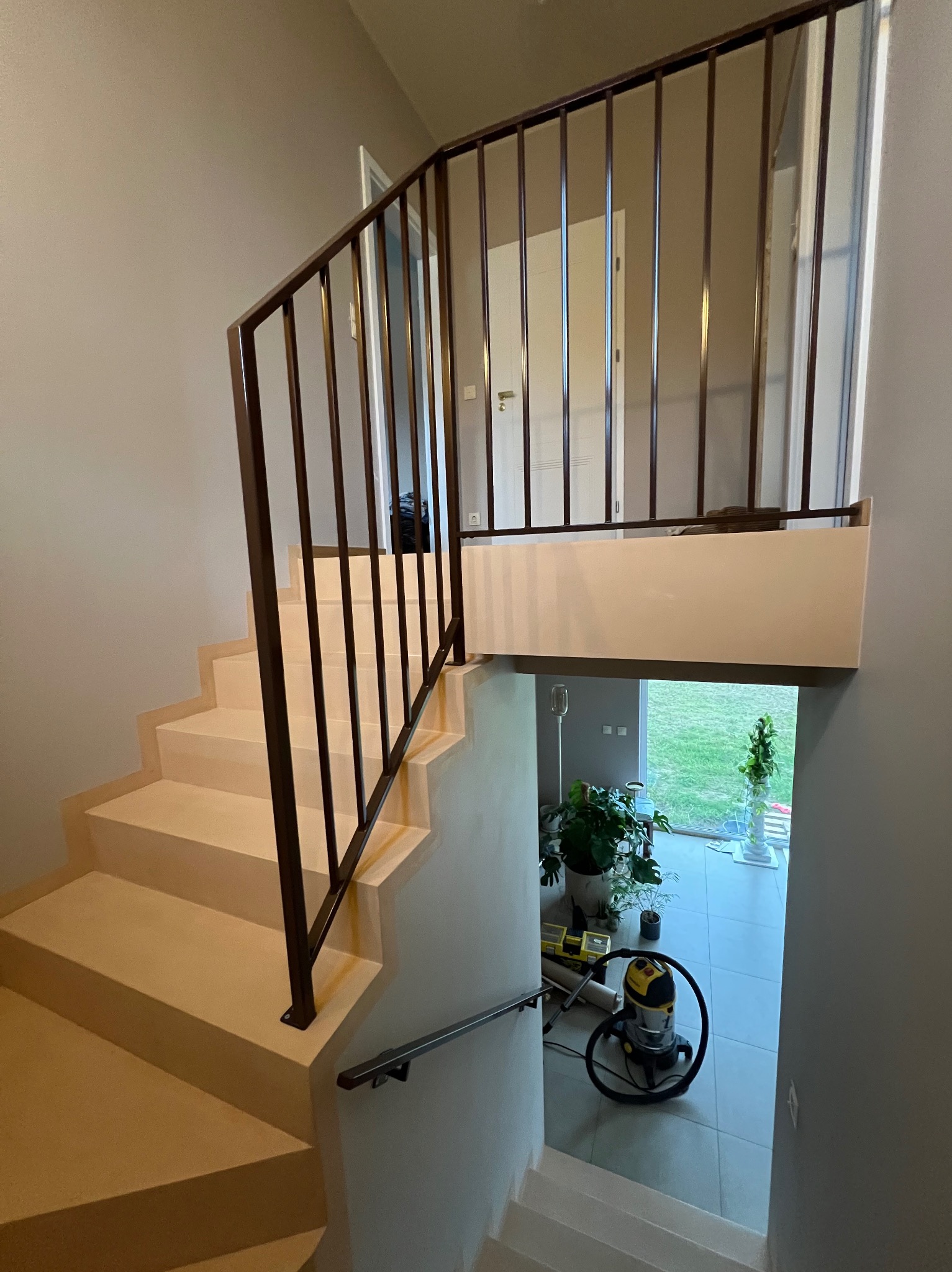 Wnętrze domu z beżowymi schodami i ciemną, metalową balustradą. Widoczny fragment poręczy i odkurzacz na dole schodów. Ujęcie z góry, z perspektywy pierwszego piętra.