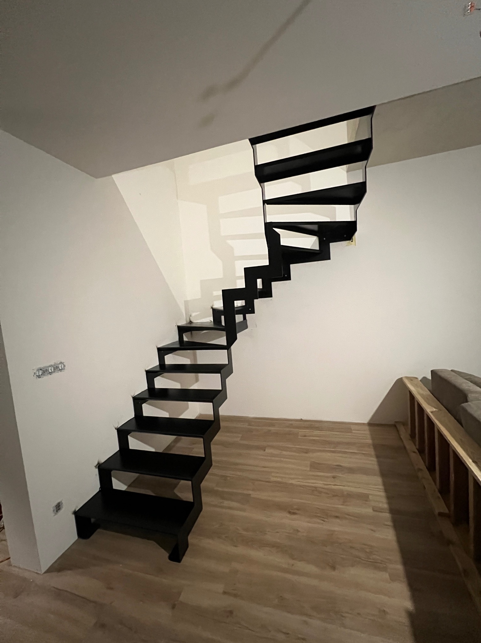 Nowoczesne, czarne schody metalowe bez balustrady, zamontowane w jasnym wnętrzu. Widoczne minimalistyczne stopnie i konstrukcja, podkreślone przez oświetlenie.