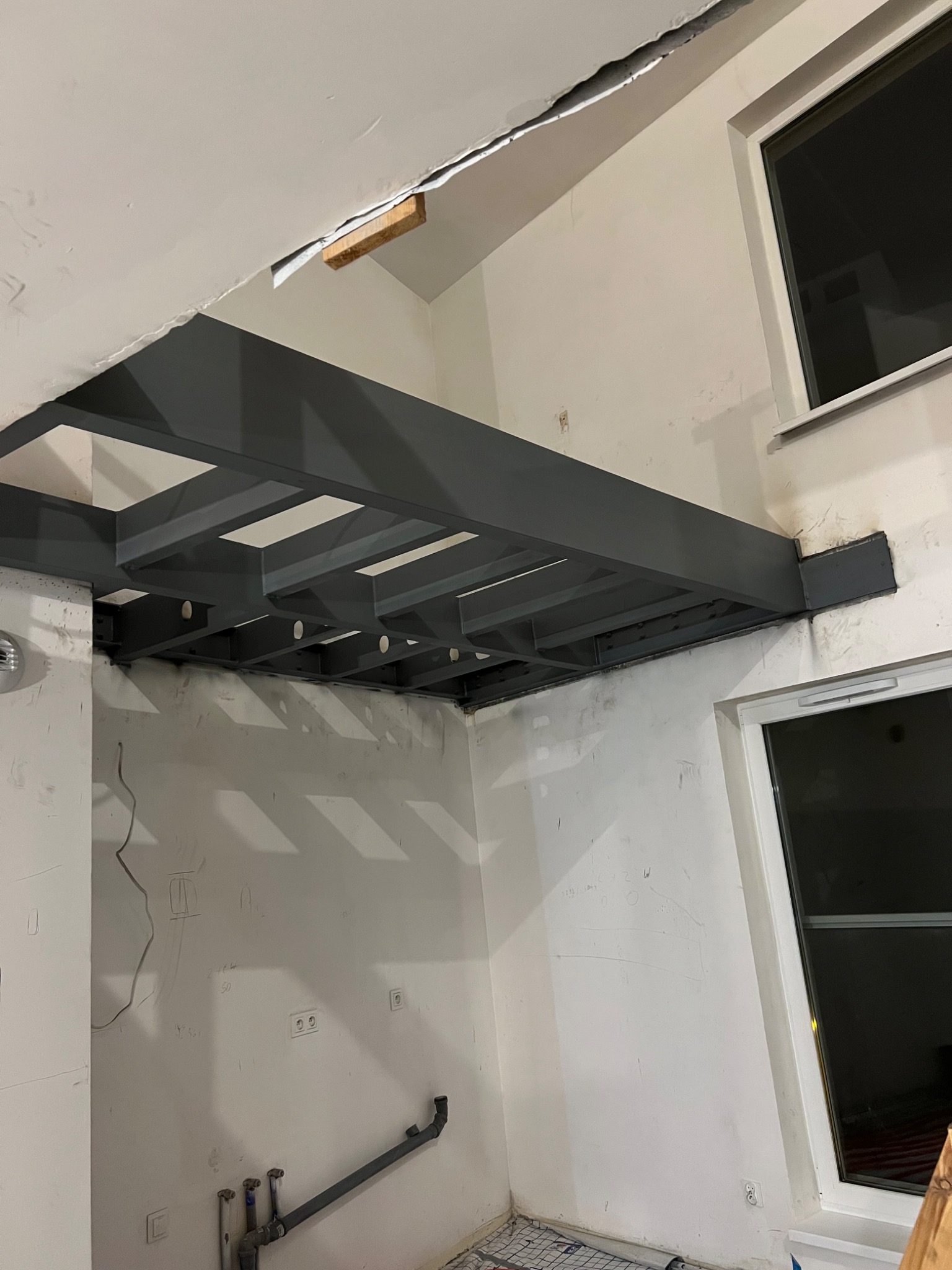 Szara, metalowa konstrukcja sufitowa z widocznymi spawami, zamontowana w surowym wnętrzu z oknami. Widoczne instalacje i niedokończone ściany.