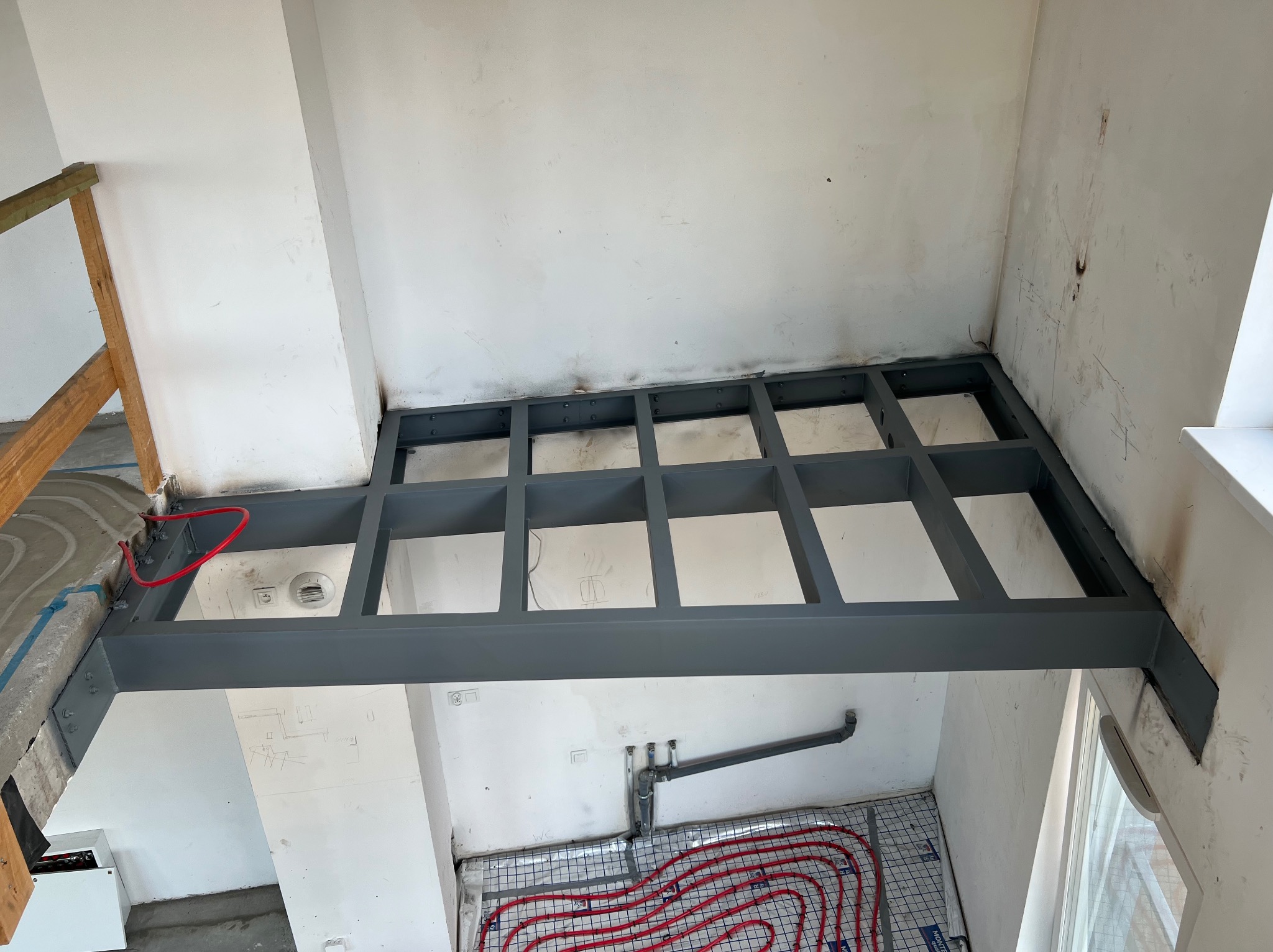 Konstrukcja metalowa pod podłogę, widok z góry na szarą kratownicę z profili, instalacja ogrzewania podłogowego w tle, surowe ściany.